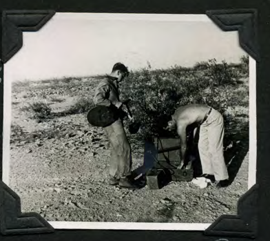 Two men assembling a mine detector on rocky desert ground.