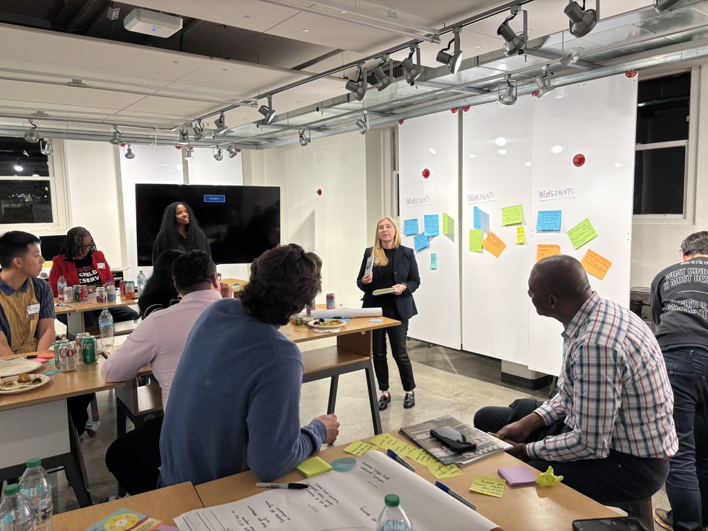 Peggy presenting to group with whiteboard showing INGREDIENTS and sticky notes