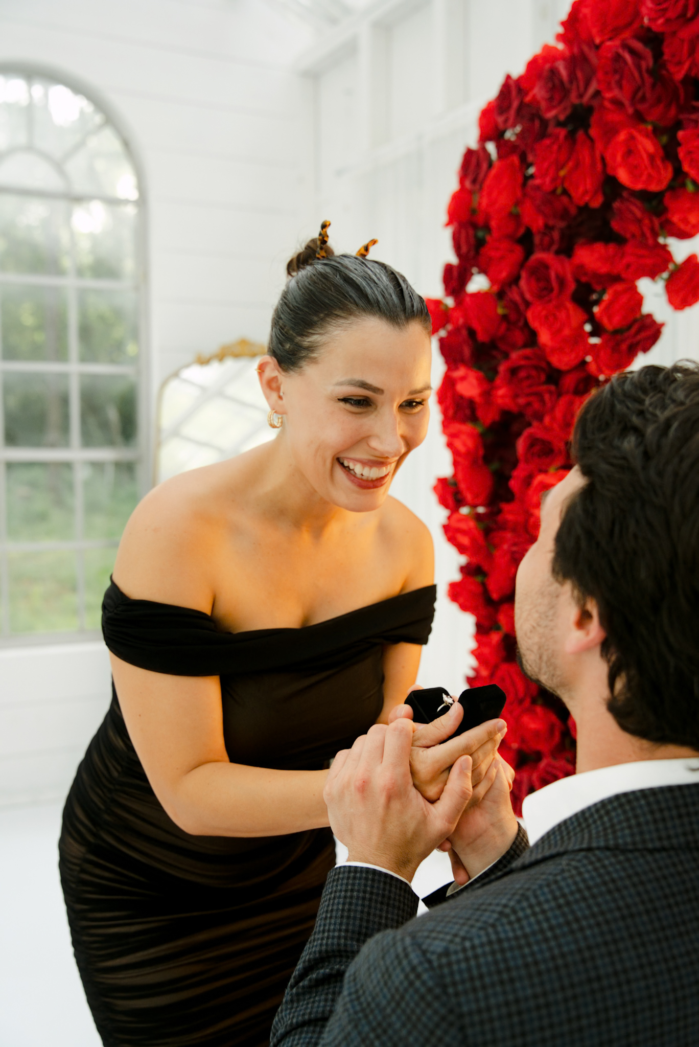 Woman in a black dress beaming as her partner presents the ring