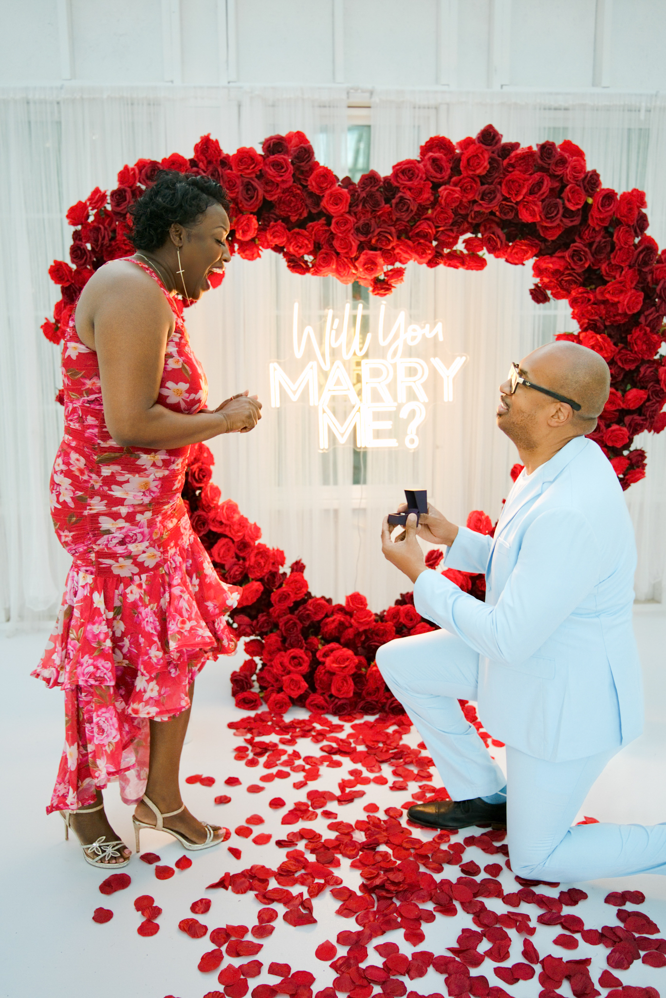 Man in powder-blue suit proposing beneath the Will You Marry Me neon in the glass house