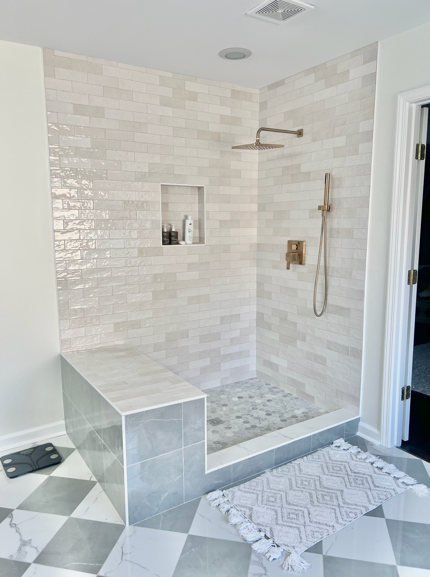 Walk-in shower with cream zellige subway tile, a tiled bench, gold fixtures, and a marble checkerboard floor