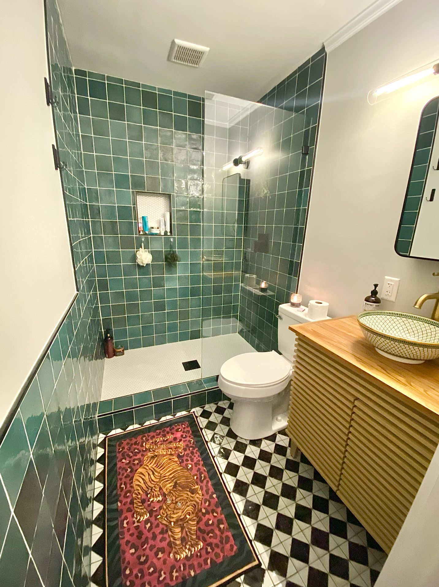 Bold green square-tile bathroom with a slatted wood vanity, glass shower, and a tiger-print rug