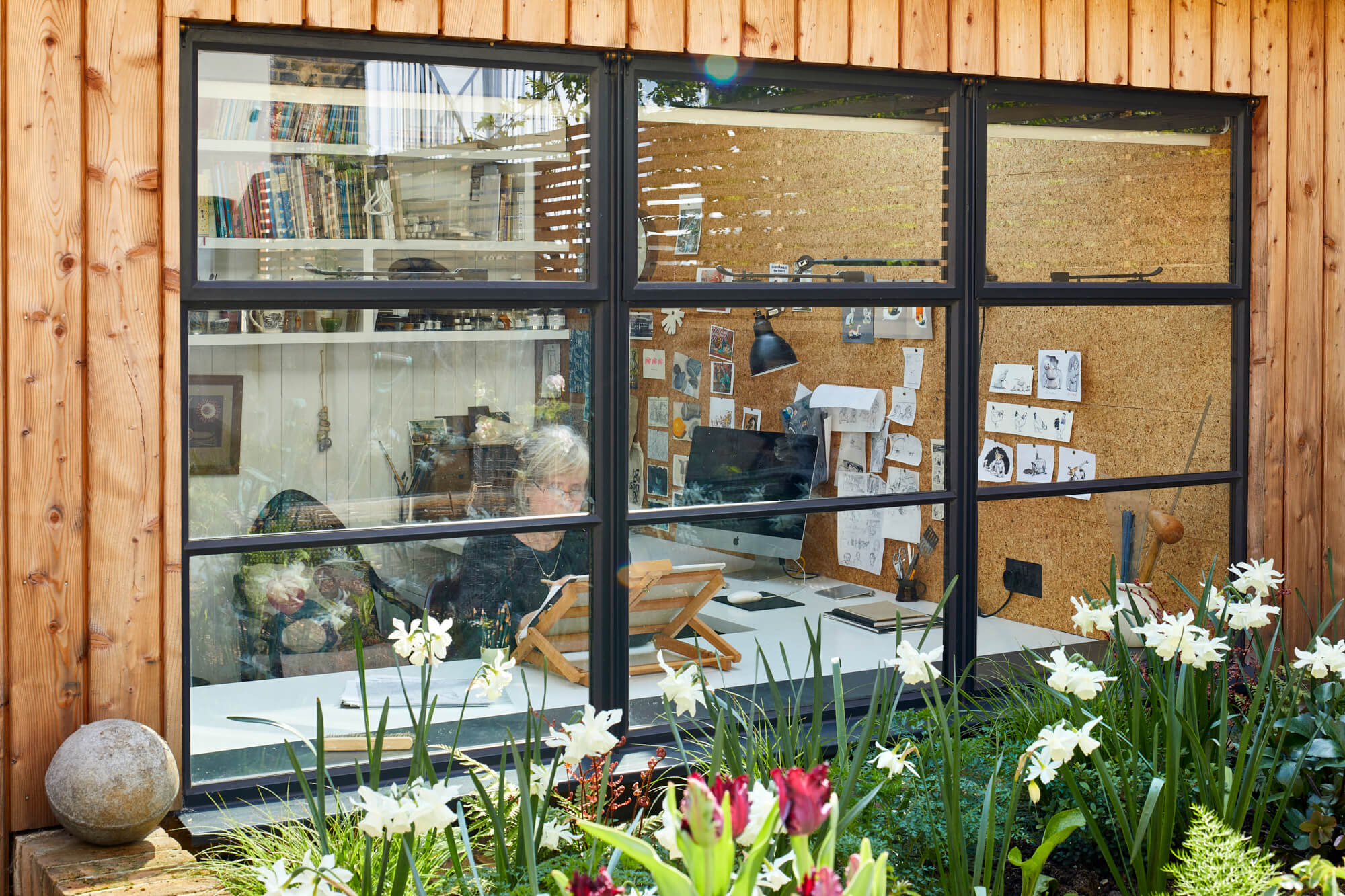 View through window showing creative workspace with cork board wall and garden beyond