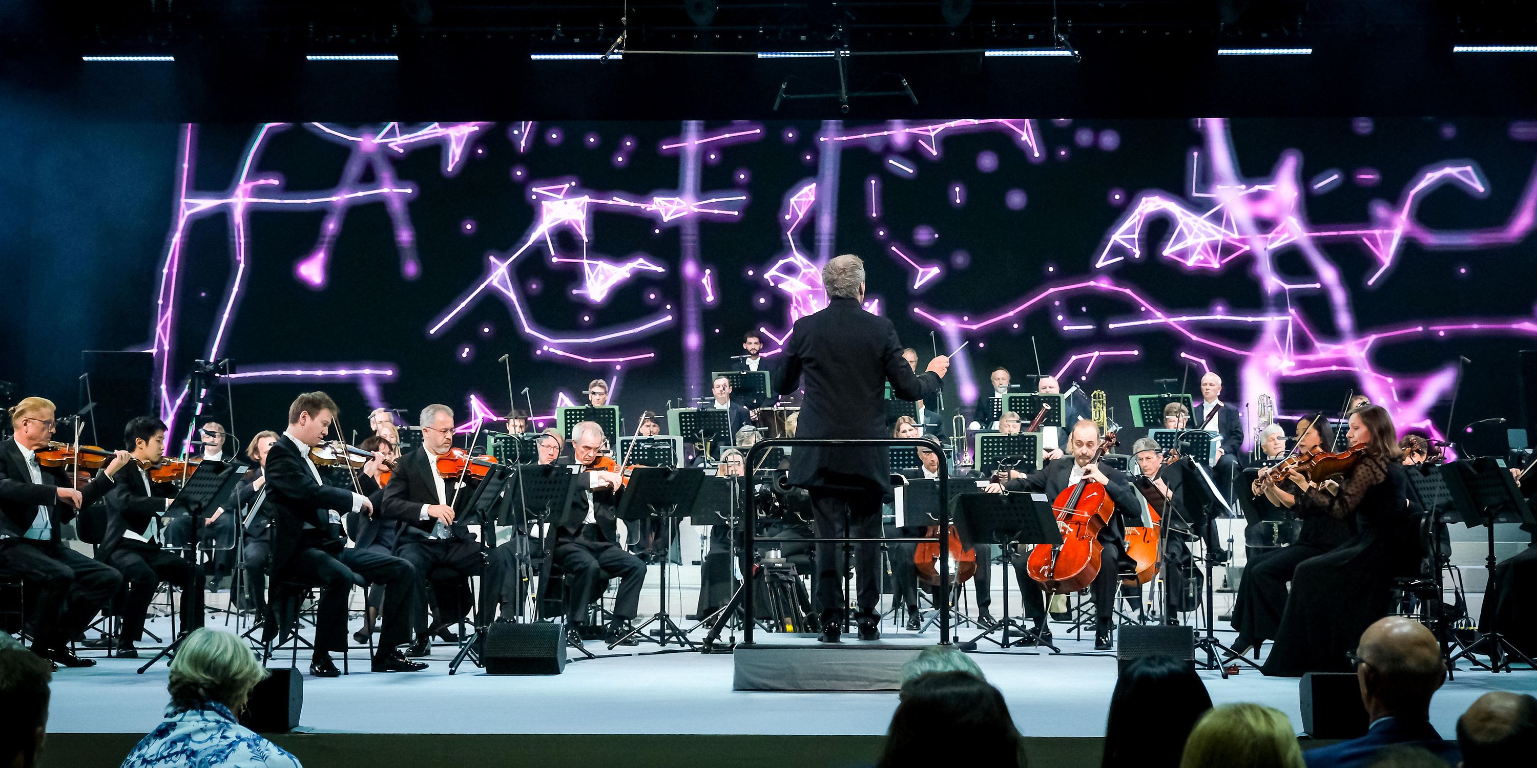Orchestra performing the Beethoven X symphony with conductor and neural network visualizations projected behind them