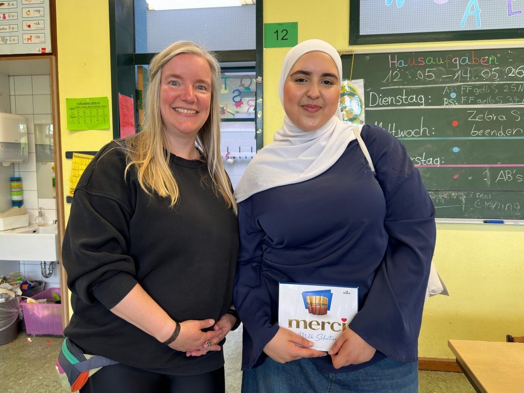 Lehrerin und Patin posieren lächelnd mit einer Merci-Schachtel im Klassenzimmer