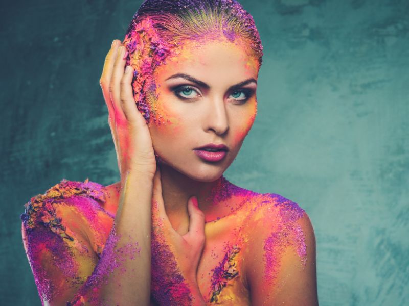 Model with vivid colorful body art and textured pigments across the face, hair, and shoulders.