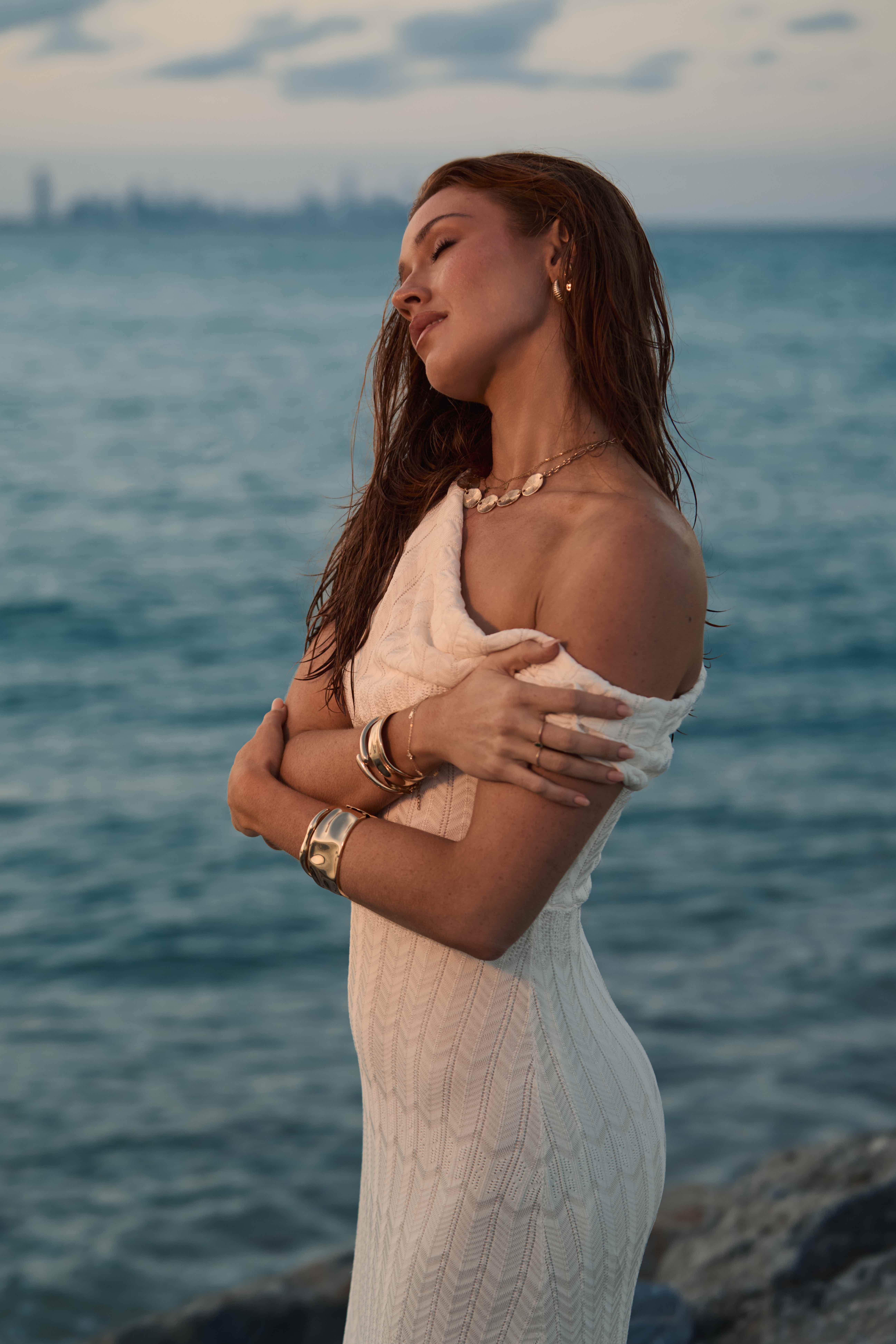 Model in white off-shoulder knit dress eyes closed by the ocean at dusk