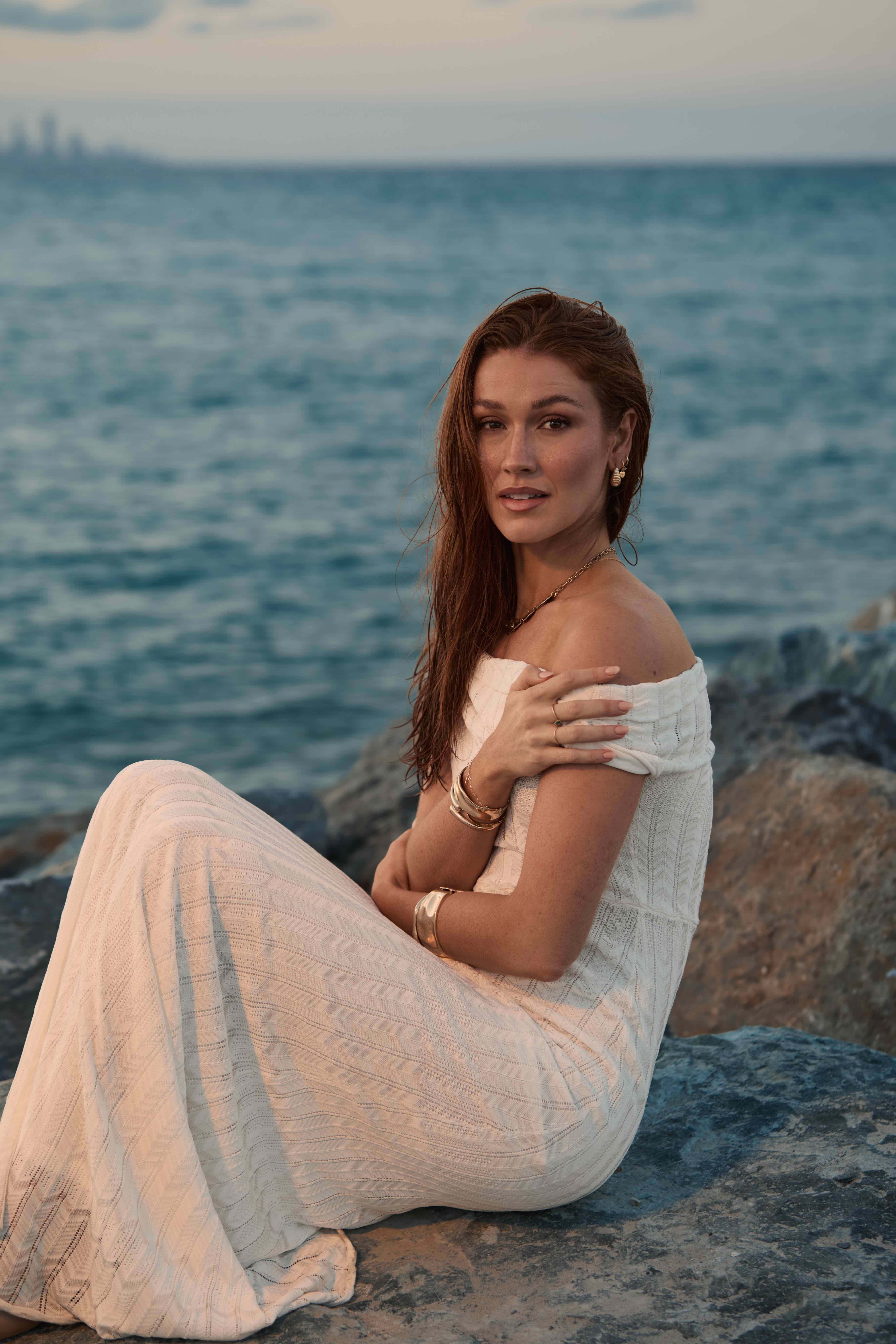 Model seated on rocks in white off-shoulder knit dress looking at camera