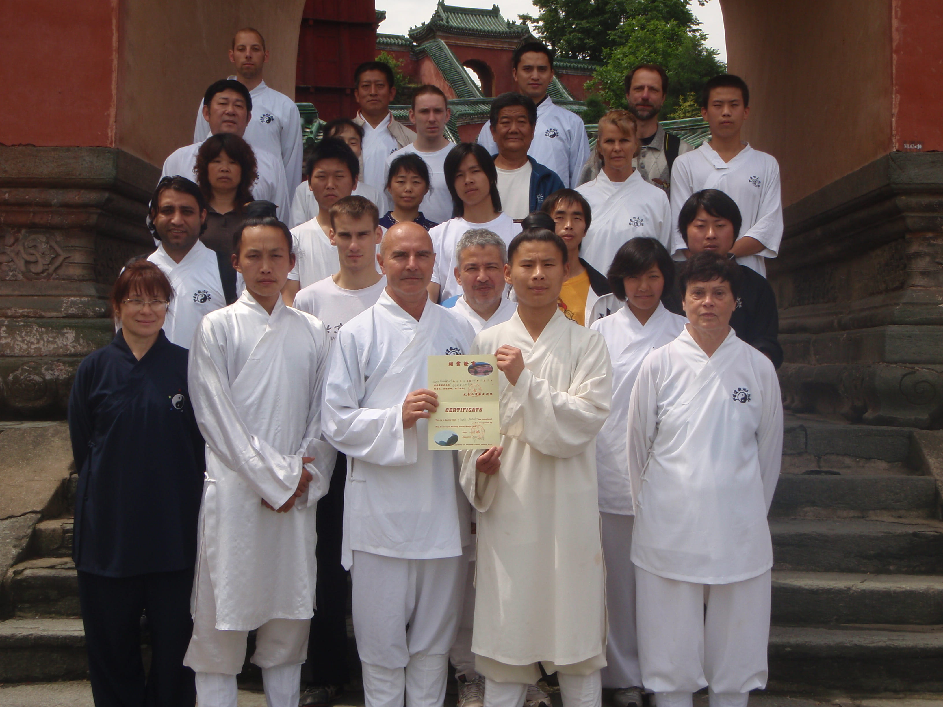 Kadama receiving his certification from a Wudang Taoist master in China, holding the official certificate alongside fellow students in white qigong robes outside a traditional temple