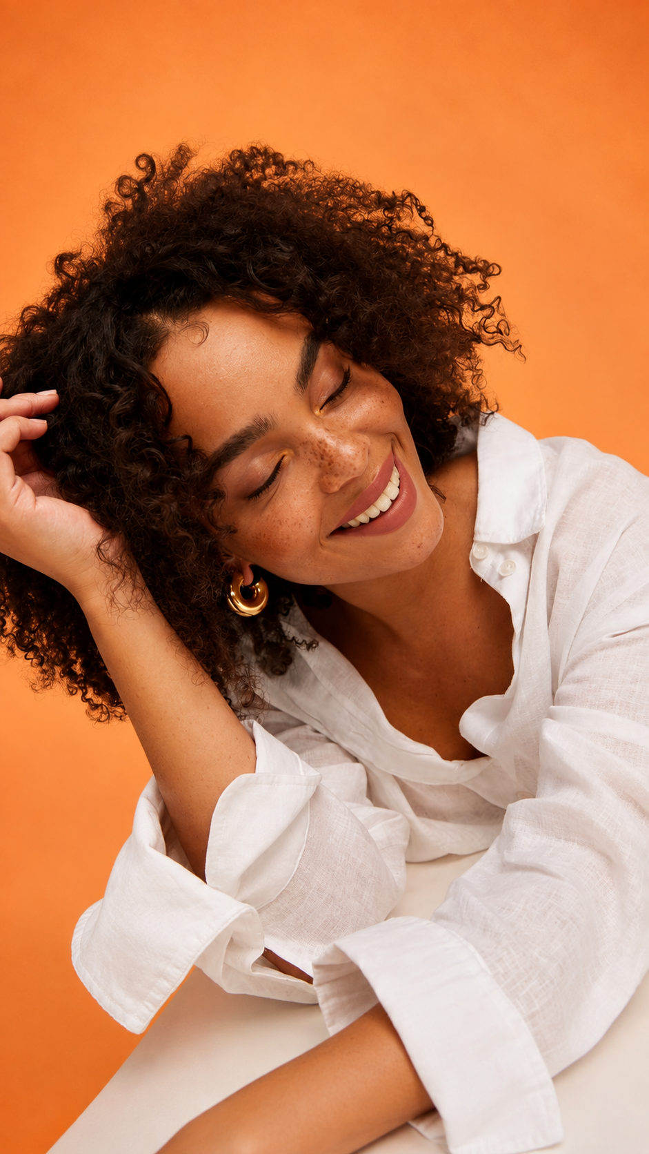 Mulher sorrindo enquanto toca os cabelos cacheados diante de um fundo laranja