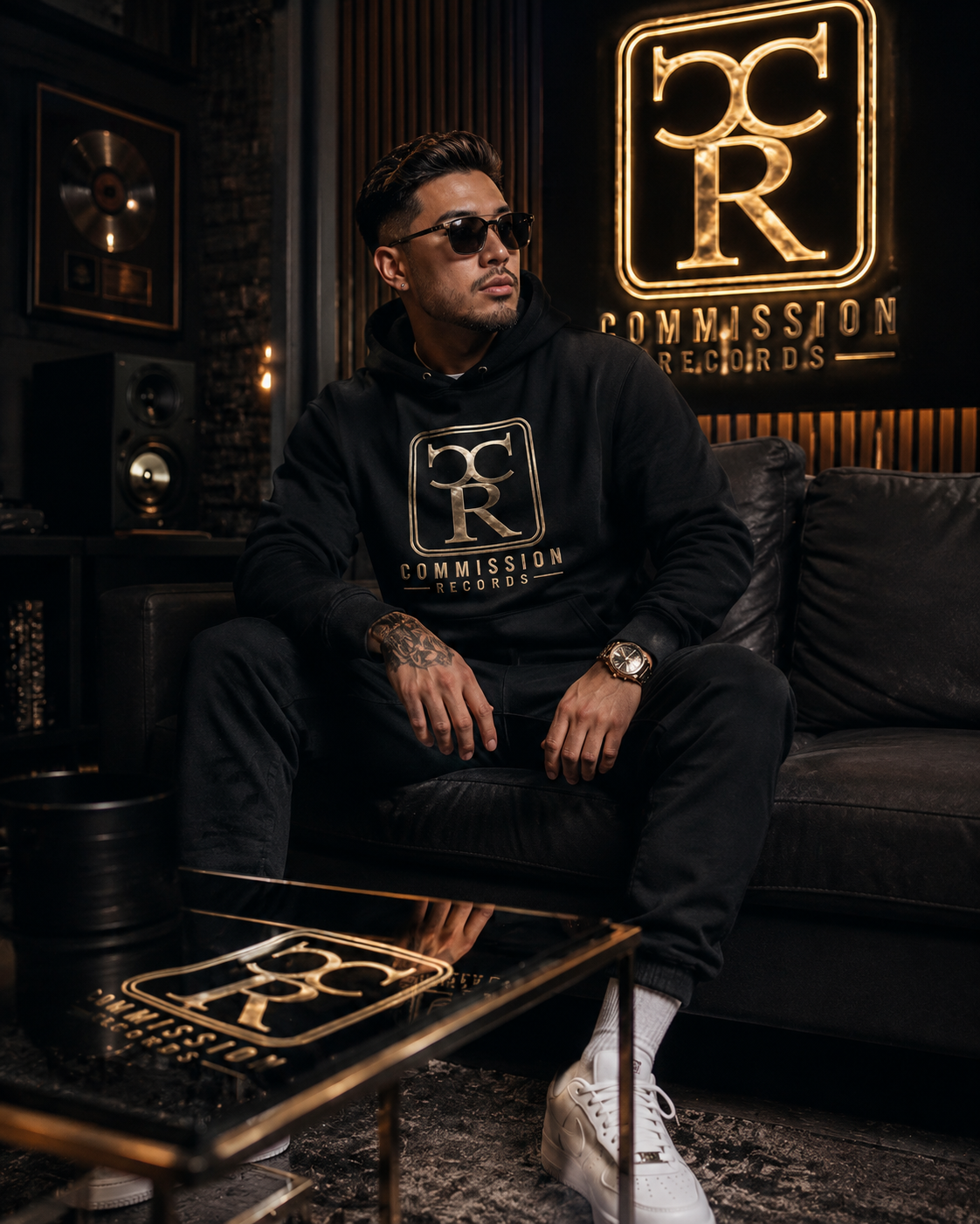 Man wearing a black Commission Records hoodie with the gold CR logo, seated in a dark studio lounge beneath a glowing gold Commission Records neon sign