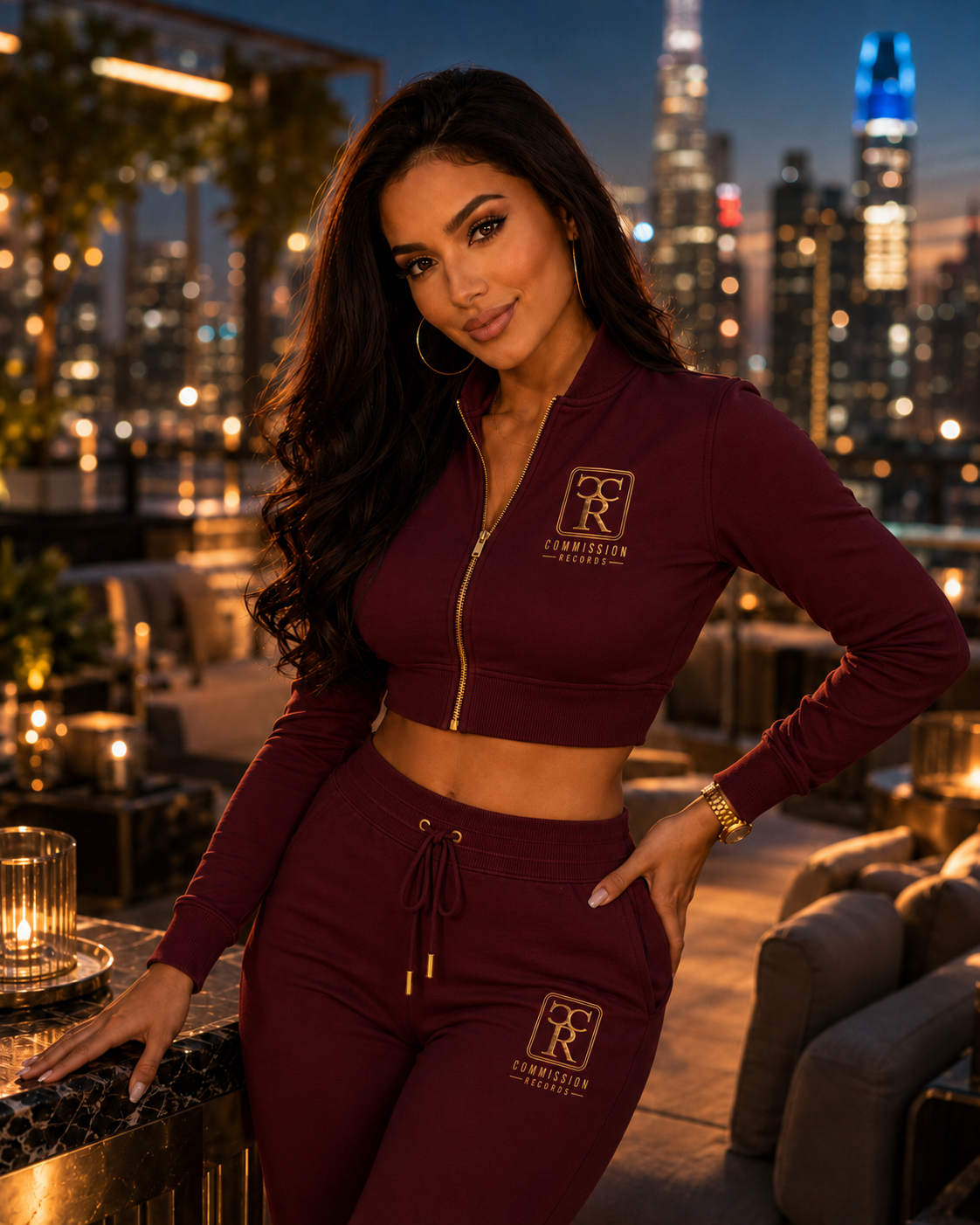 Woman in a burgundy cropped zip Commission Records track jacket and matching joggers, posing on a rooftop lounge at dusk