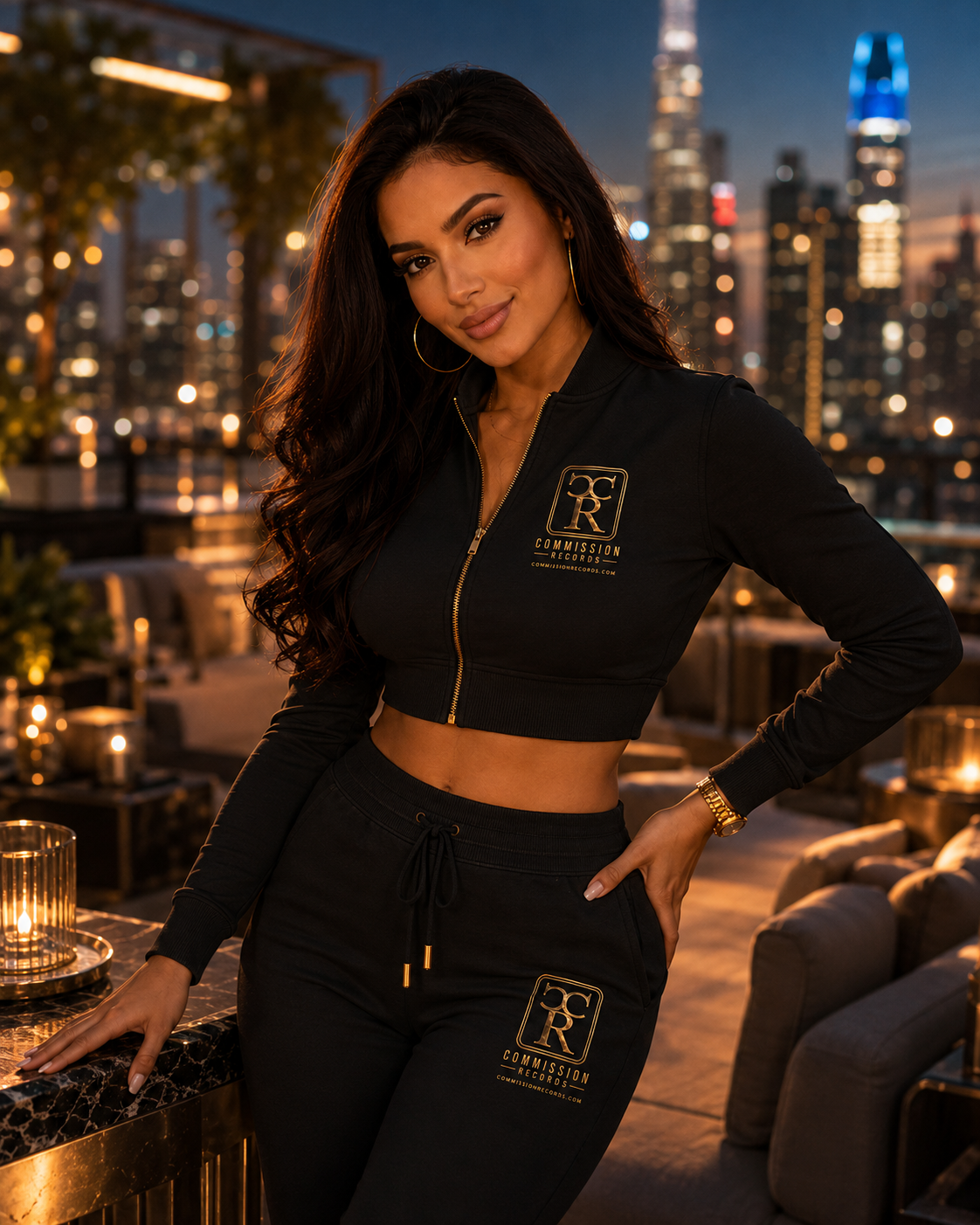 Woman in a black cropped zip Commission Records track jacket and matching joggers, posing on a rooftop lounge with a city skyline behind her
