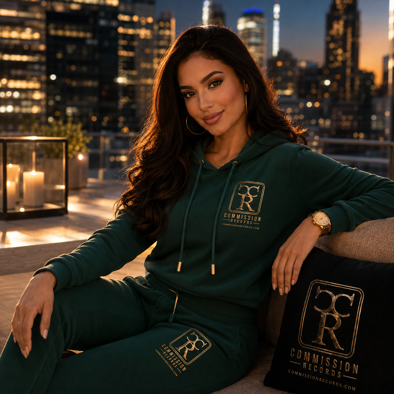 Woman in an emerald green Commission Records pullover hoodie and matching sweatpants, seated on a rooftop couch at sunset beside a black CR logo pillow