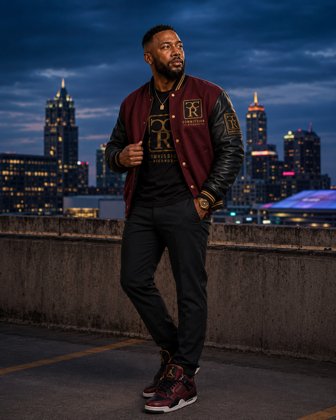 Man on a rooftop at dusk wearing a burgundy-and-black leather-sleeve Commission Records varsity jacket over a black-and-gold CR logo tee with black pants and burgundy Jordan 4 sneakers
