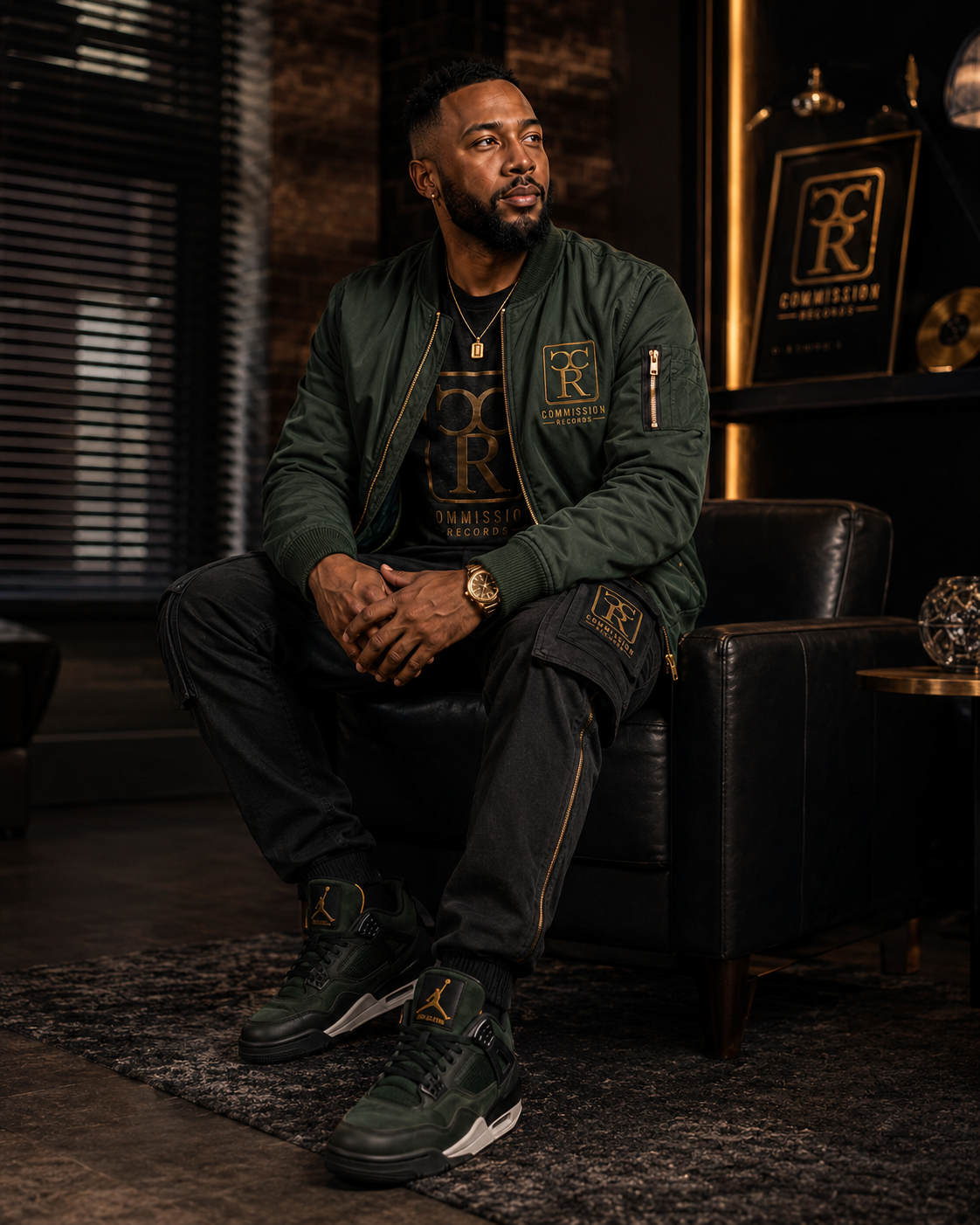 Man seated in a lounge wearing an olive green Commission Records bomber jacket over a black-and-gold CR logo tee with black CR cargo pants and green-and-gold Jordan 4 sneakers