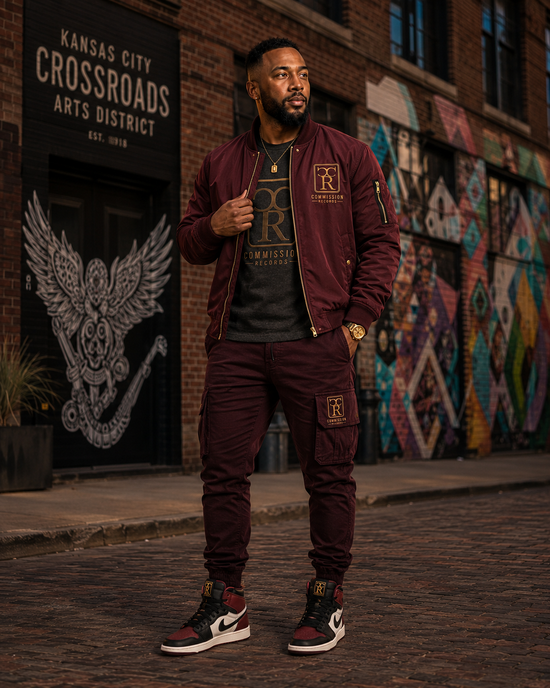 Man in the Kansas City Crossroads Arts District wearing a burgundy Commission Records bomber jacket over a CR logo tee with matching burgundy CR cargo joggers and burgundy Jordan 1 sneakers