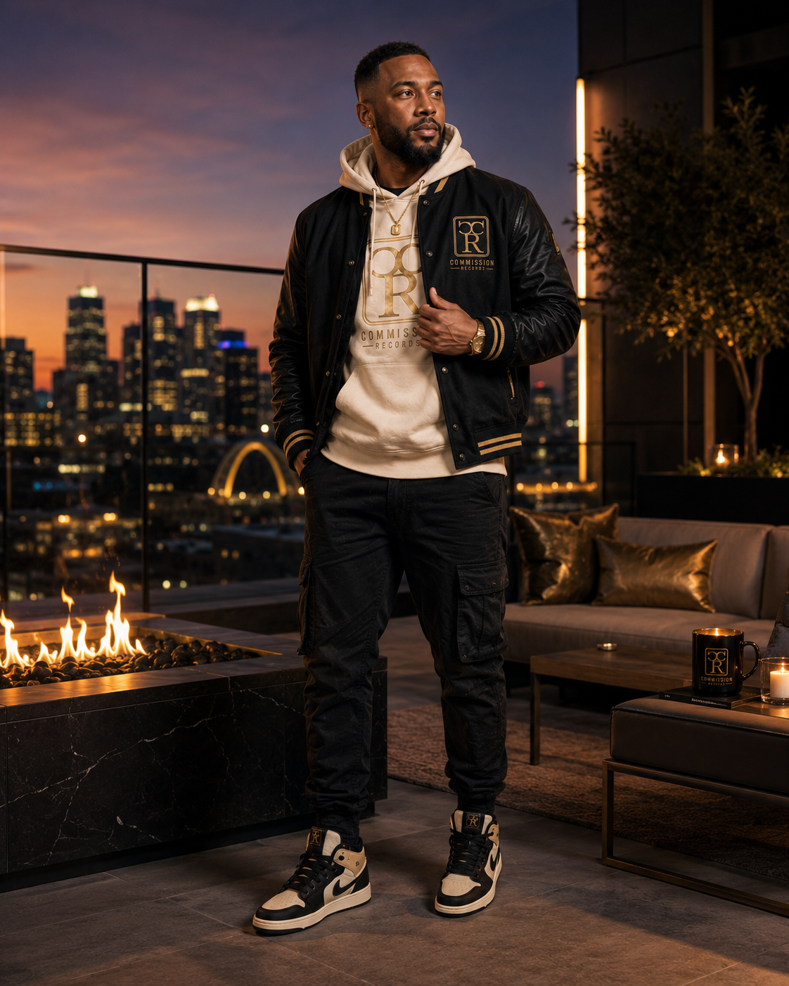 Man on a rooftop terrace by a firepit wearing a black-and-gold leather-sleeve Commission Records varsity jacket over a cream CR hoodie with black cargo pants and cream-and-black Jordan 1 sneakers