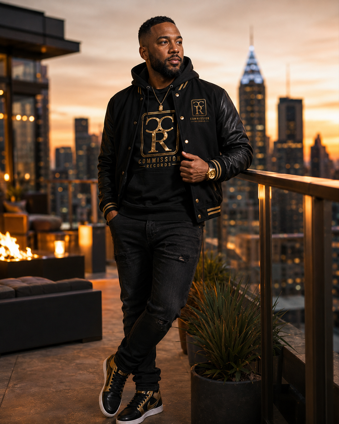 Man on a rooftop at sunset wearing a black-and-gold leather-sleeve Commission Records varsity jacket over a black-and-gold CR logo tee with black ripped jeans and black-and-gold Jordan 1 sneakers
