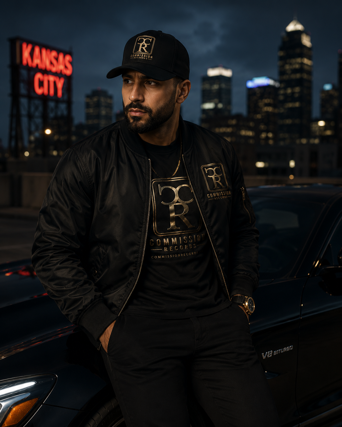 Man leaning on a black car by a Kansas City neon sign, wearing a black-and-gold Commission Records CR logo snapback cap with a black bomber jacket over a black-and-gold CR tee