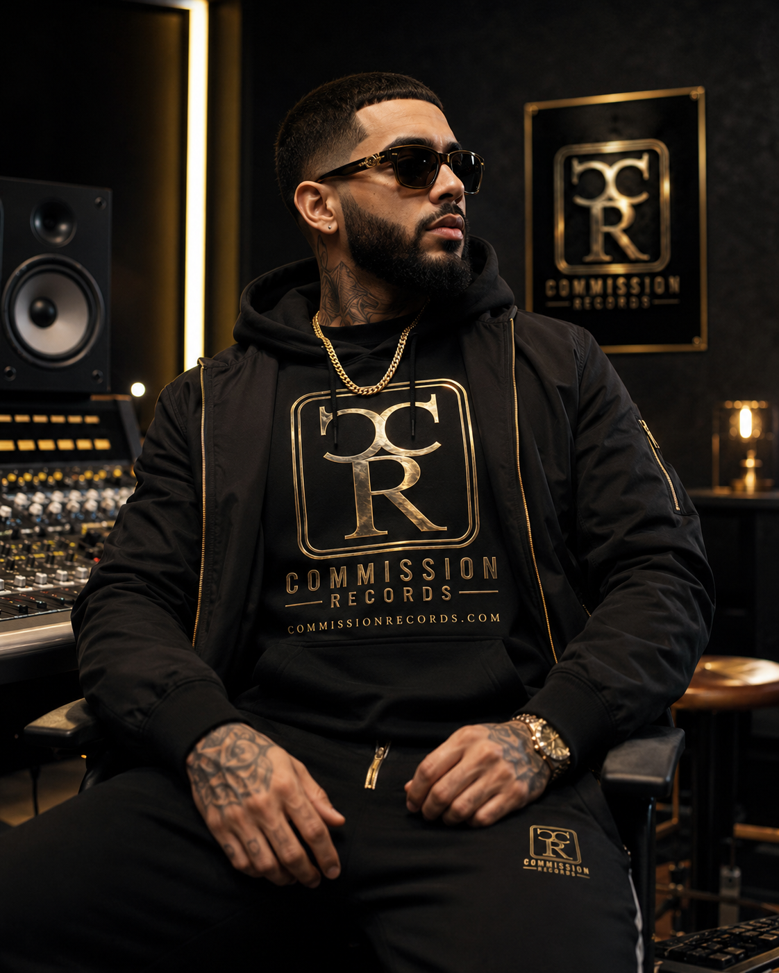 Man in sunglasses wearing a black-and-gold Commission Records hoodie and matching sweatpants under a black bomber jacket, seated in a studio beside a gold CR logo poster