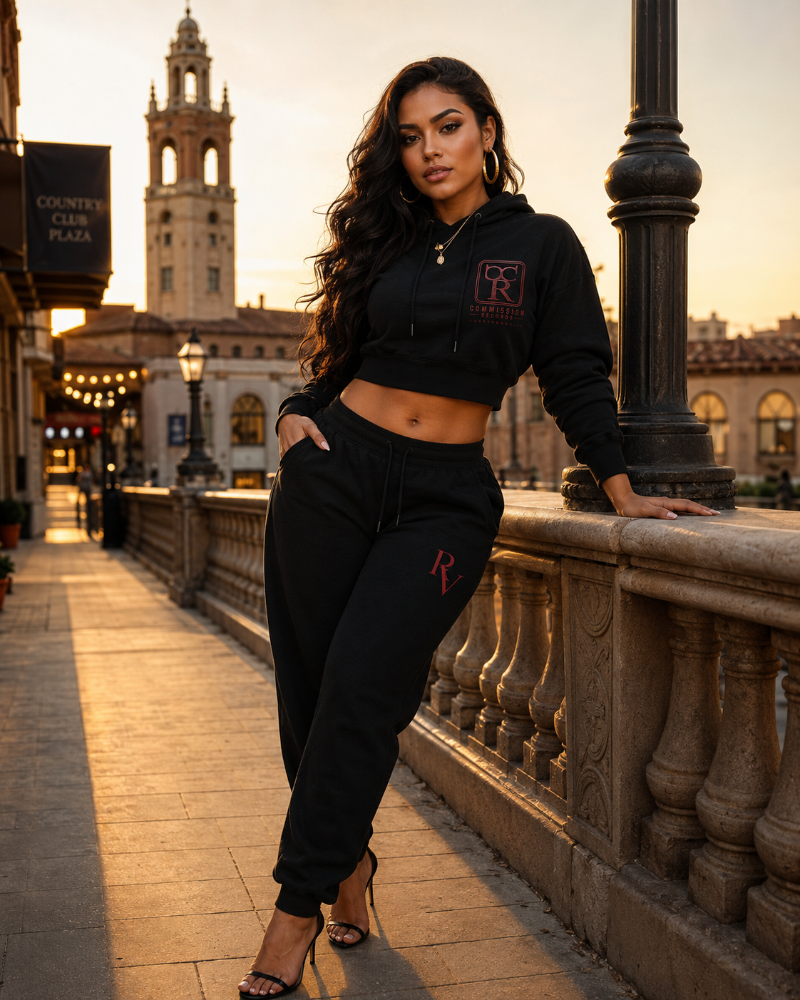 Woman on the Country Club Plaza at sunset, wearing a black cropped hoodie with a red Commission Records CR logo and matching black Rome Valentino RV joggers with heels, leaning on a stone balustrade