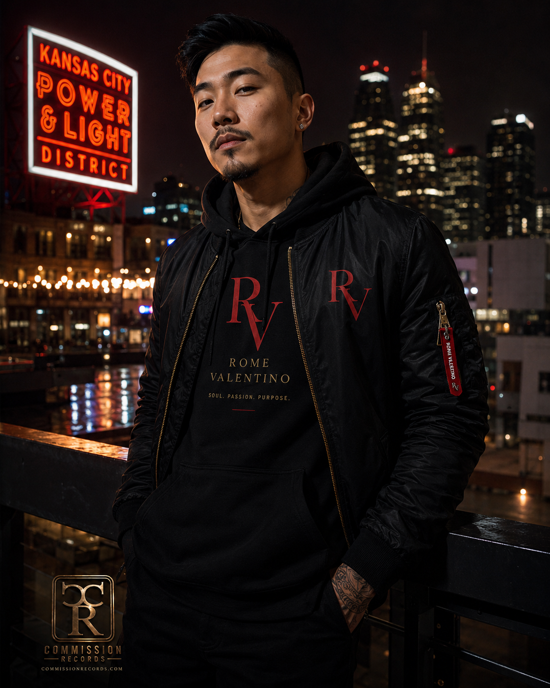 Man on a rooftop by the Kansas City Power & Light District neon sign, wearing a black bomber jacket with a red Rome Valentino RV zipper pull over a black Rome Valentino RV hoodie with Soul Passion Purpose print