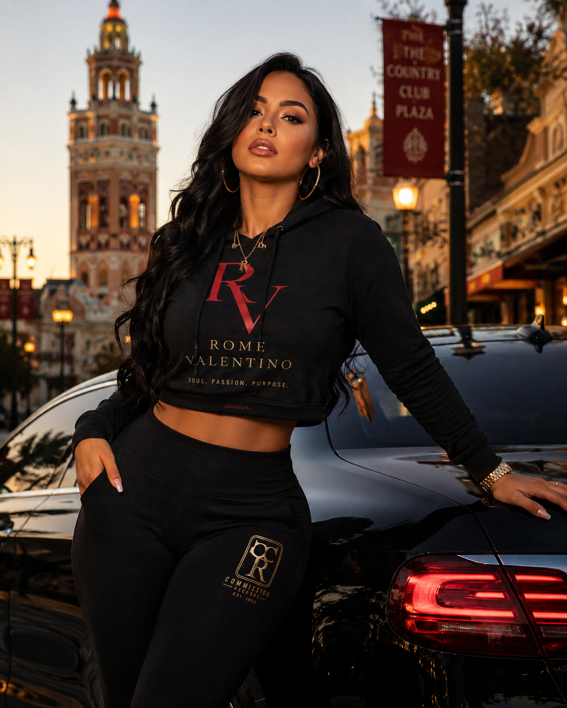 Woman on the Country Club Plaza at sunset, wearing a black Rome Valentino RV cropped hoodie with red and gold Soul Passion Purpose print and matching black joggers with a gold Commission Records CR logo, leaning on a black car