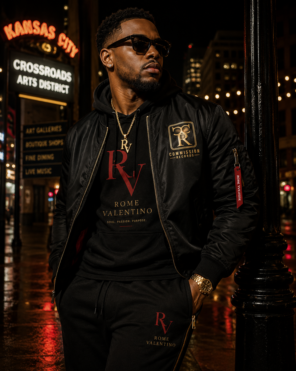 Man in sunglasses on a wet Kansas City Crossroads Arts District street at night, wearing a black bomber jacket with a gold Commission Records CR patch over a black Rome Valentino RV hoodie with Soul Passion Purpose print and matching black RV joggers