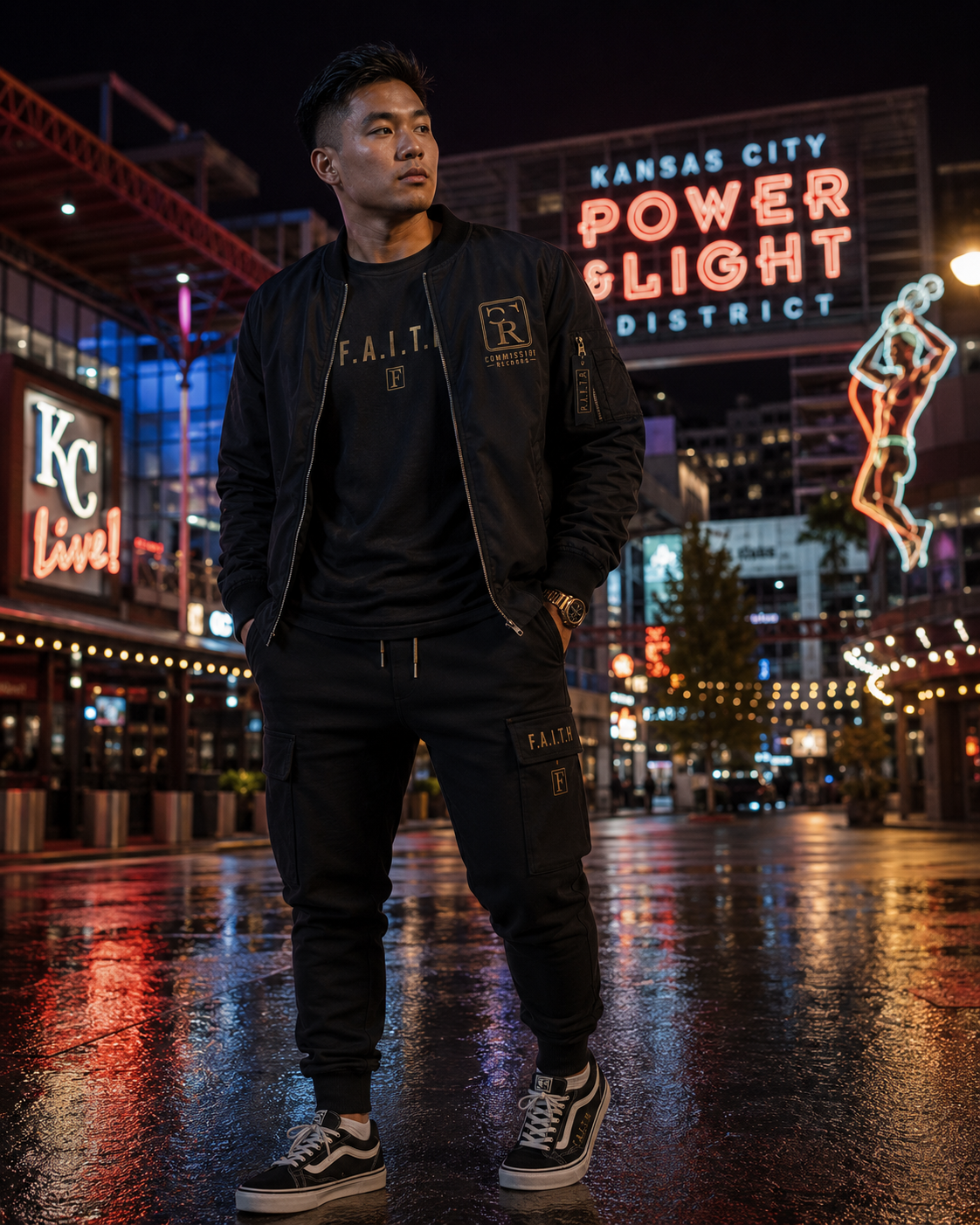 Man in the Kansas City Power & Light District on a wet plaza at night, wearing a black bomber jacket with a gold Commission Records CR patch and F.A.I.T.H. sleeve tab over a black F.A.I.T.H. tee with matching black F.A.I.T.H. cargo joggers