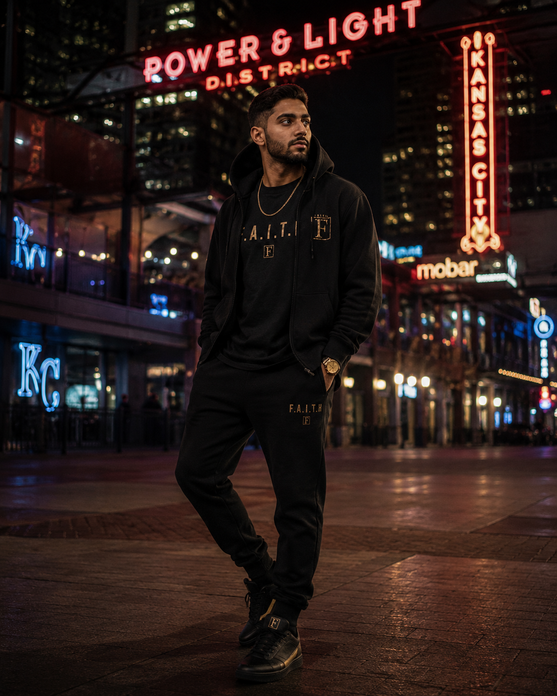 Man in the Kansas City Power & Light District at night wearing a full black F.A.I.T.H. zip-up hoodie and jogger tracksuit set with gold boxed-F accents and gold-trimmed sneakers