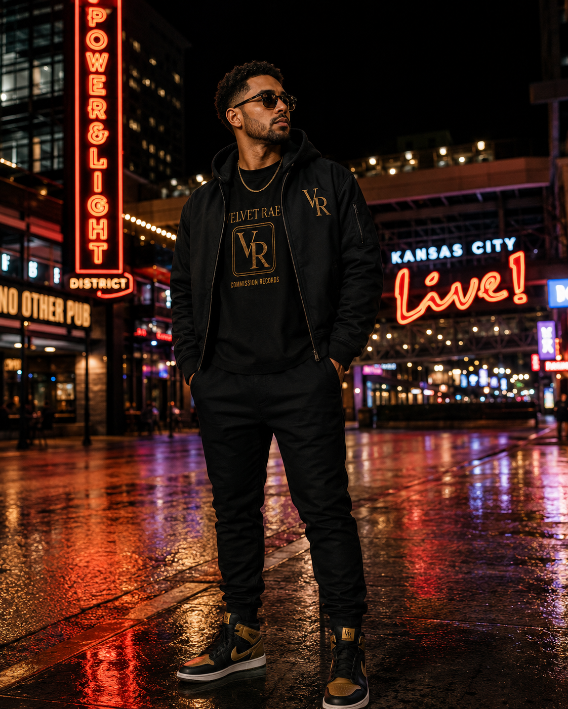 Man in the Kansas City Power & Light District at night wearing a black hooded bomber over a black Velvet Rae VR crest tee, with black joggers and black-and-gold VR high-top sneakers