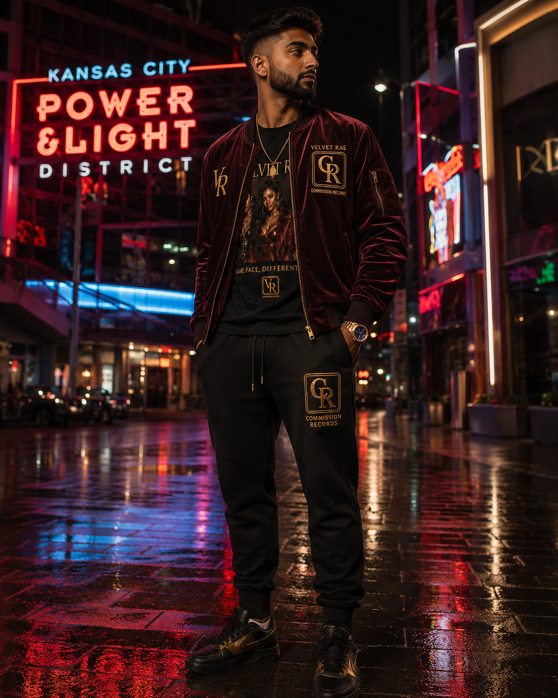Man in the Kansas City Power & Light District at night wearing a burgundy velvet Velvet Rae VR bomber with a gold Commission Records CR chest crest over a Same Face Different Love VR graphic tee, with black CR gold-logo joggers