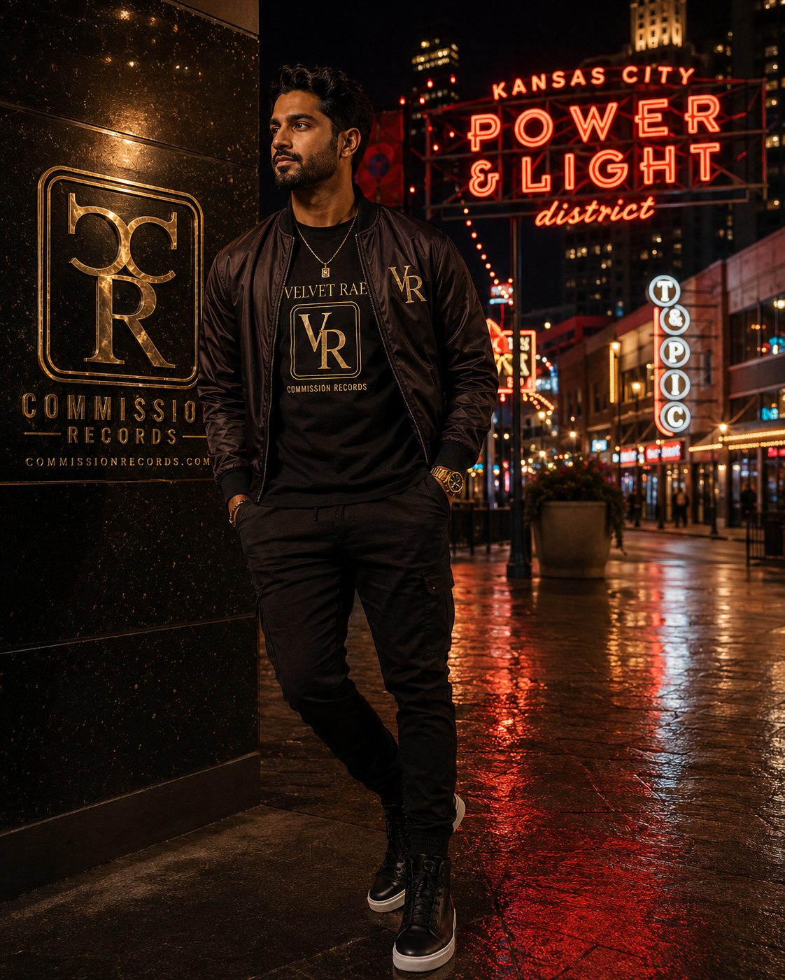 Man leaning on a gold Commission Records CR plaque wall in the Kansas City Power & Light District at night, wearing a black bomber jacket with a gold VR sleeve logo over a black Velvet Rae VR crest tee with black cargo joggers