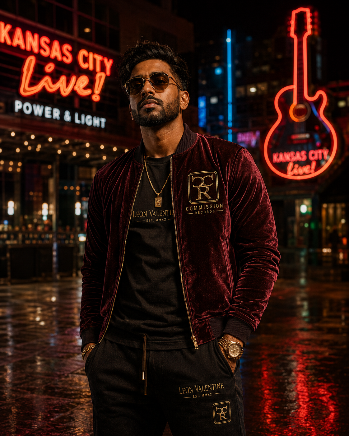 Man in the Kansas City Power & Light District at night, wearing a burgundy velvet Commission Records bomber jacket with a gold CR chest crest over a black Leon Valentine EST. MMXX tee, with matching black Leon Valentine CR joggers