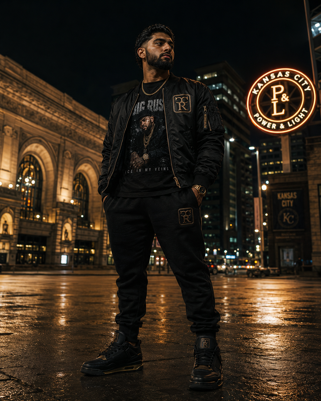 Man at Kansas City Union Station beneath the Power & Light neon sign at night wearing a black satin CR bomber with gold trim over a Big Rust Ice In My Veins portrait graphic tee, with matching black CR joggers and black-and-gold CR sneakers