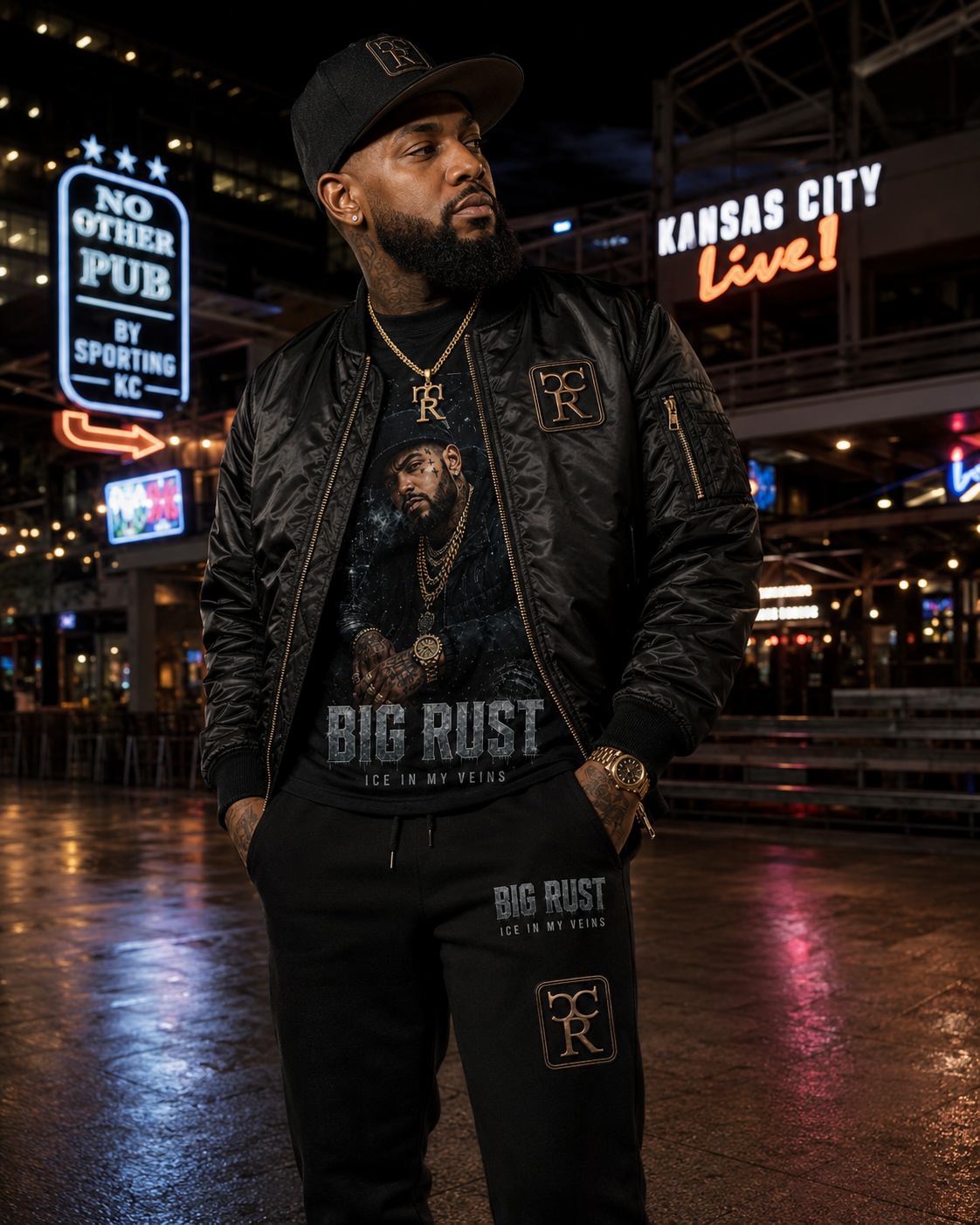 Man in the Kansas City Power & Light District at night wearing a black satin CR bomber with gold trim over a Big Rust Ice In My Veins portrait graphic tee, with matching black CR joggers and a black CR fitted cap