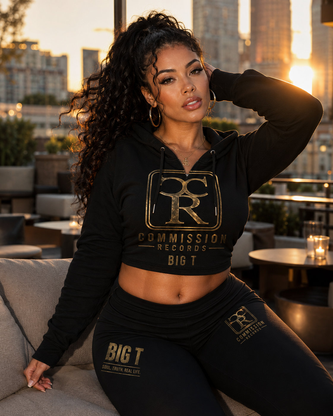 Woman on a rooftop lounge at sunset wearing a black Commission Records Big T cropped hoodie with a gold CR crest, paired with matching black CR and Big T Soul Truth Real Life leggings