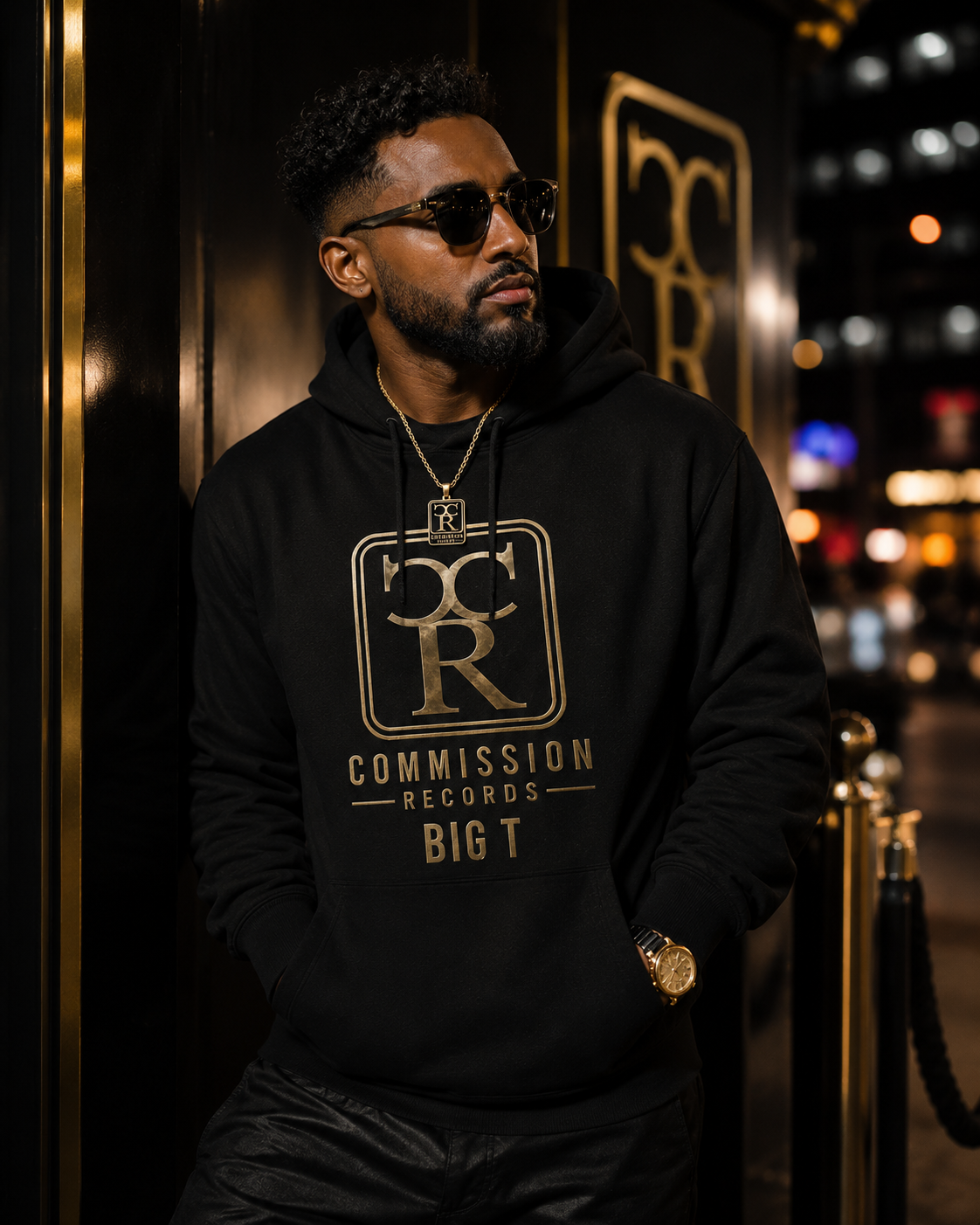 Man leaning against a gold-accented wall on a city street at night wearing a black Commission Records Big T pullover hoodie with a gold CR crest, sunglasses, and a gold CR pendant