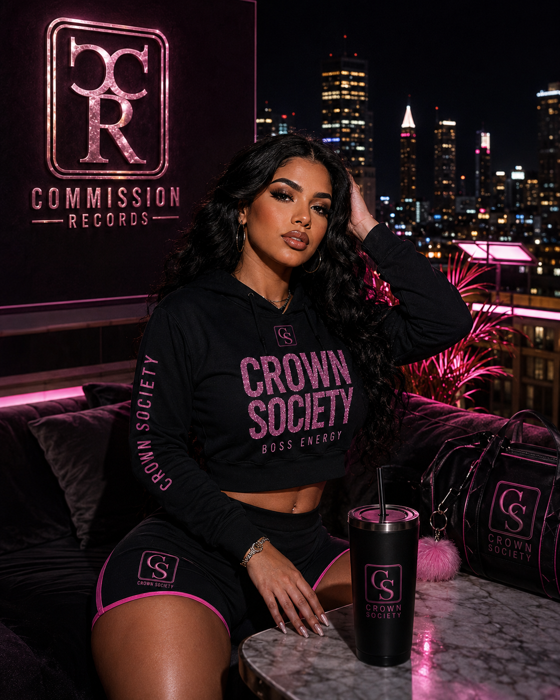 Woman on a rooftop at night wearing a black Crown Society Boss Energy pink-glitter cropped hoodie with a CS sleeve print, matching black CS pink-trim biker shorts, with a CS tumbler and CS duffel bag