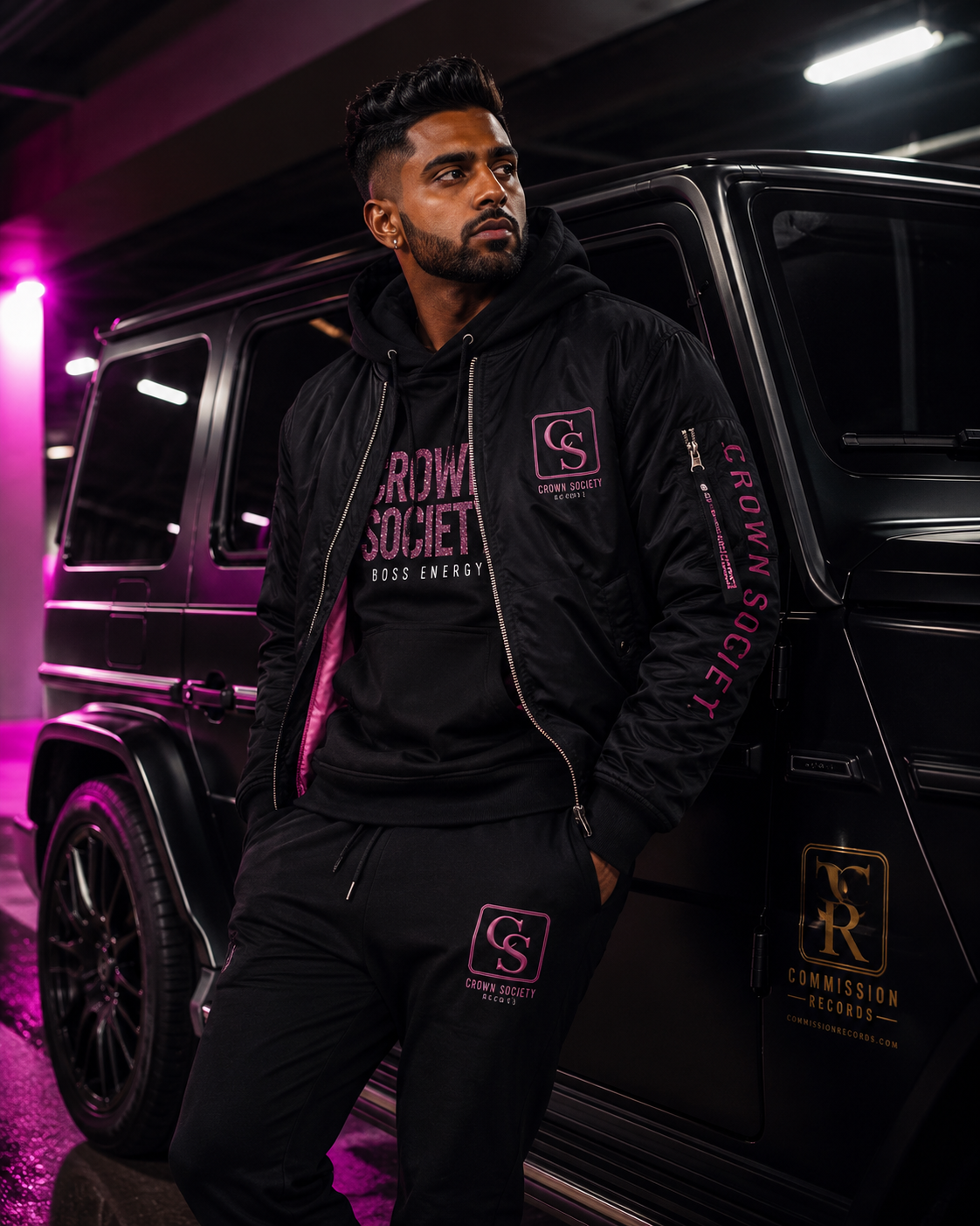 Man in a parking garage beside a black G-Wagon, wearing a black Crown Society Boss Energy pink-glitter hoodie under a black CS bomber with pink accents and a Crown Society sleeve print, with matching black CS joggers