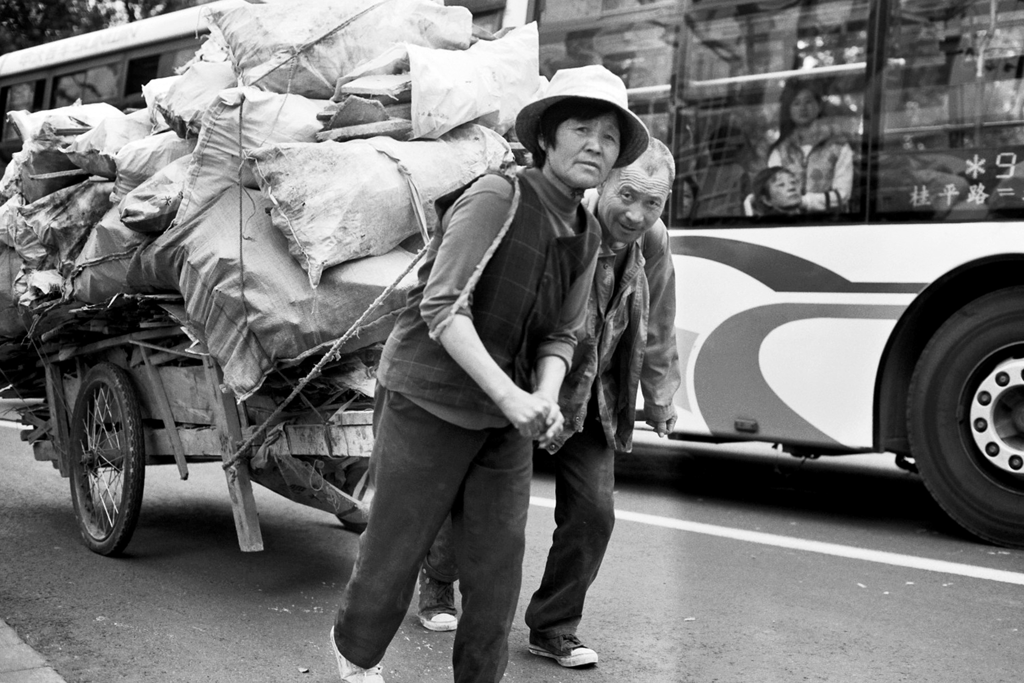 Woman pulling heavily loaded cart of recycling sacks past city bus with passengers