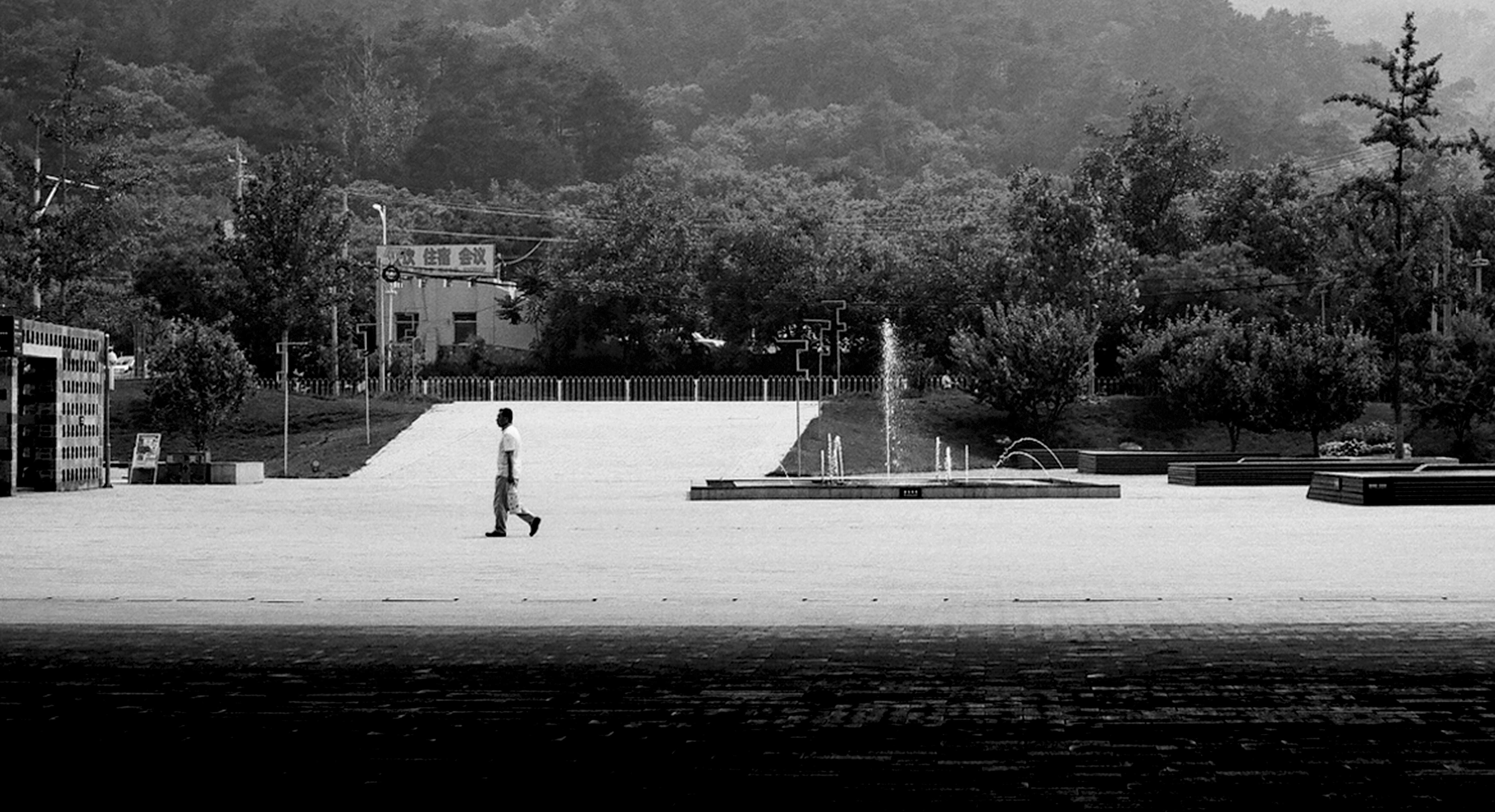 Solitary figure walking across vast empty plaza with fountain and forested mountains beyond