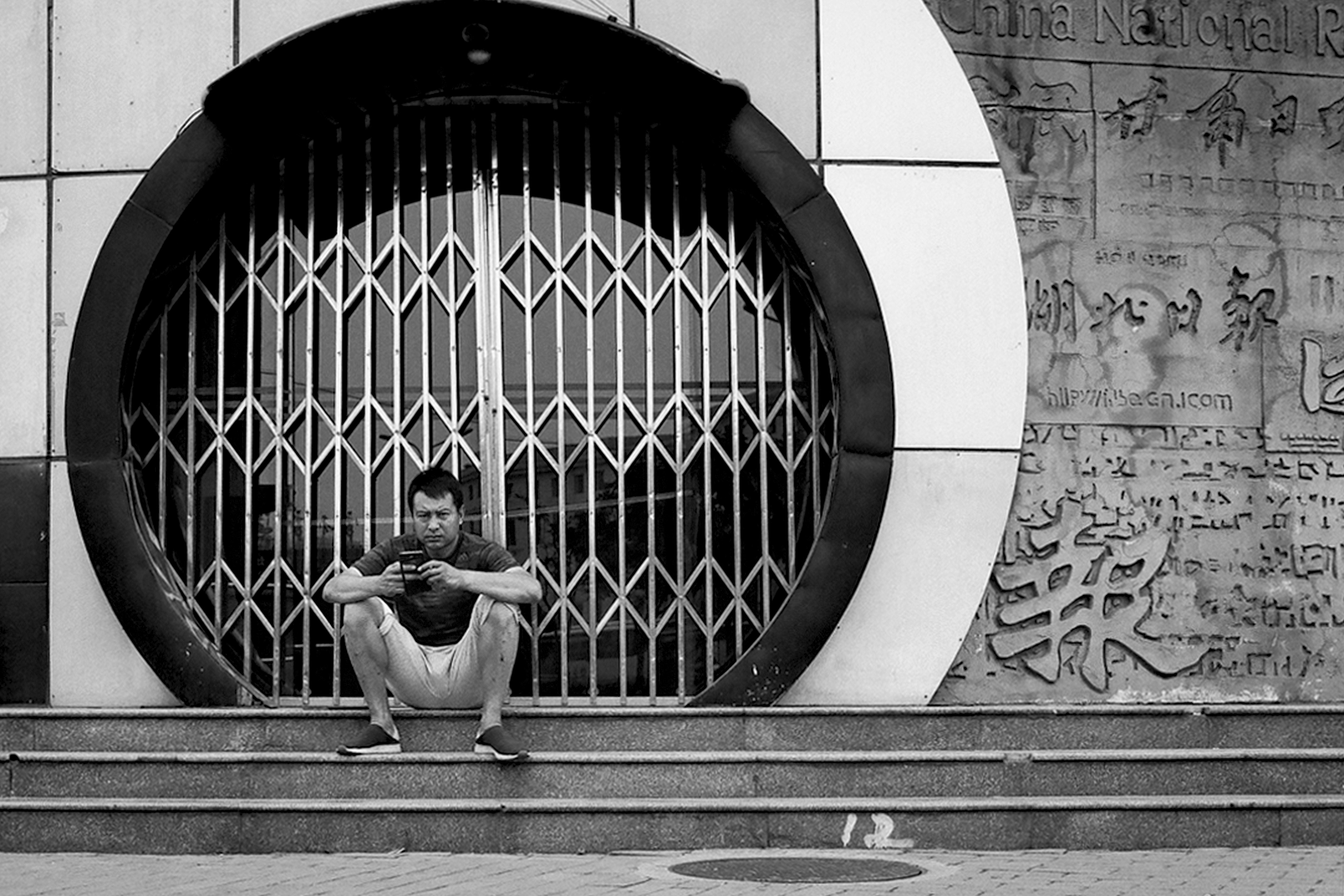 Man sitting on steps within circular moon gate entrance of building with metal lattice gate