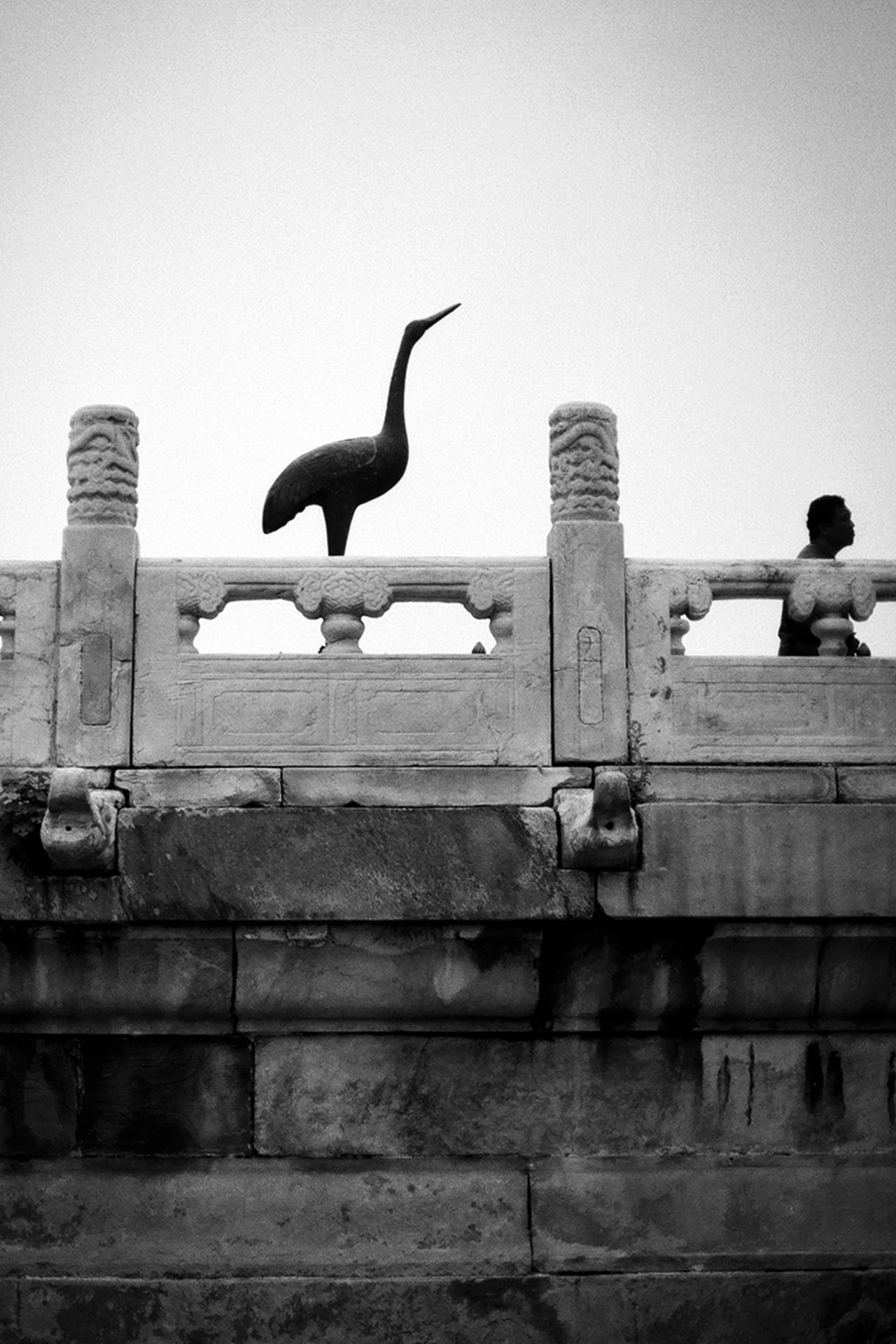 Ceramic crane sculpture standing on stone balustrade overlooking water with figure visible in background