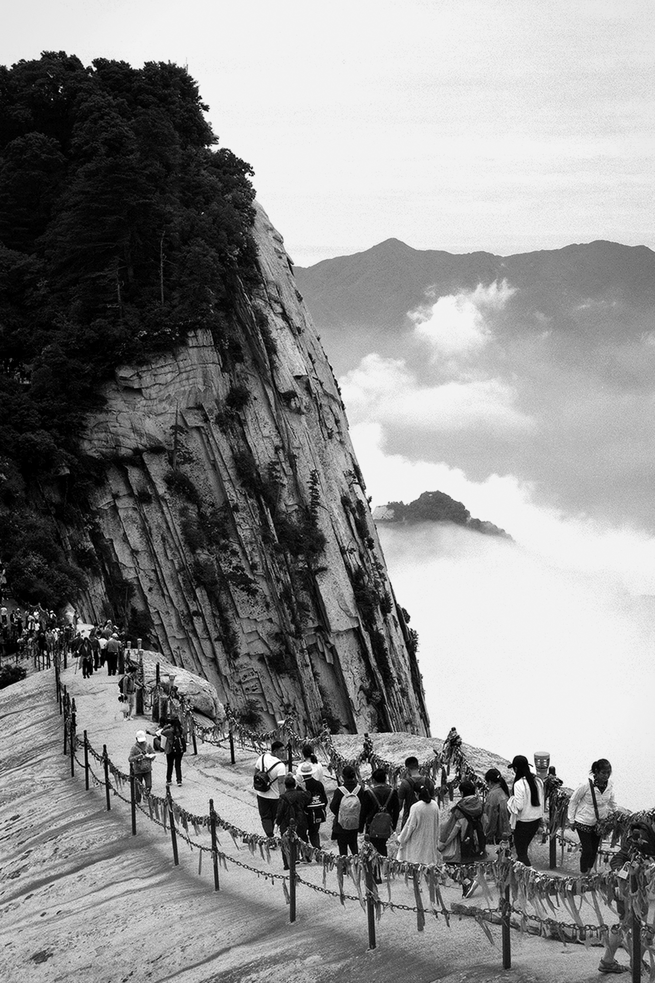 Mountain cliff pathway with visitors walking along rope railing overlooking misty forested valleys