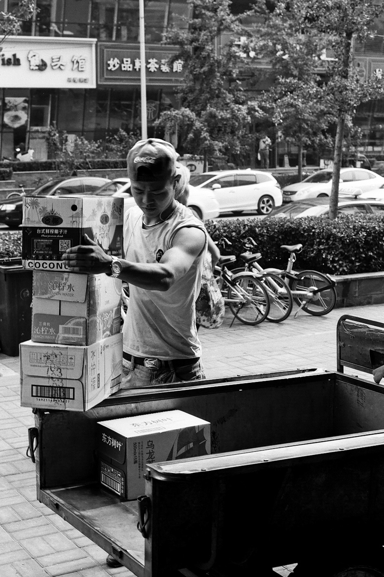 Woman loading boxes into truck bed in urban shopping area with parked bicycles and vegetation