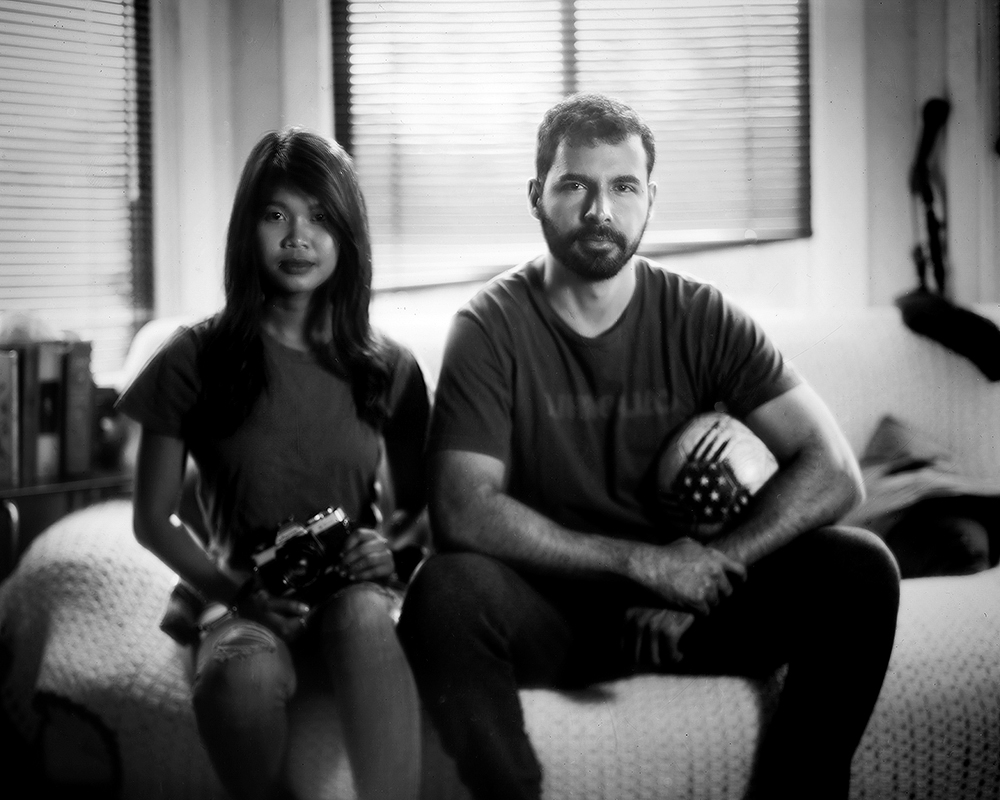 Two people sitting together in interior space with venetian blinds, woman holding camera and man with decorative object
