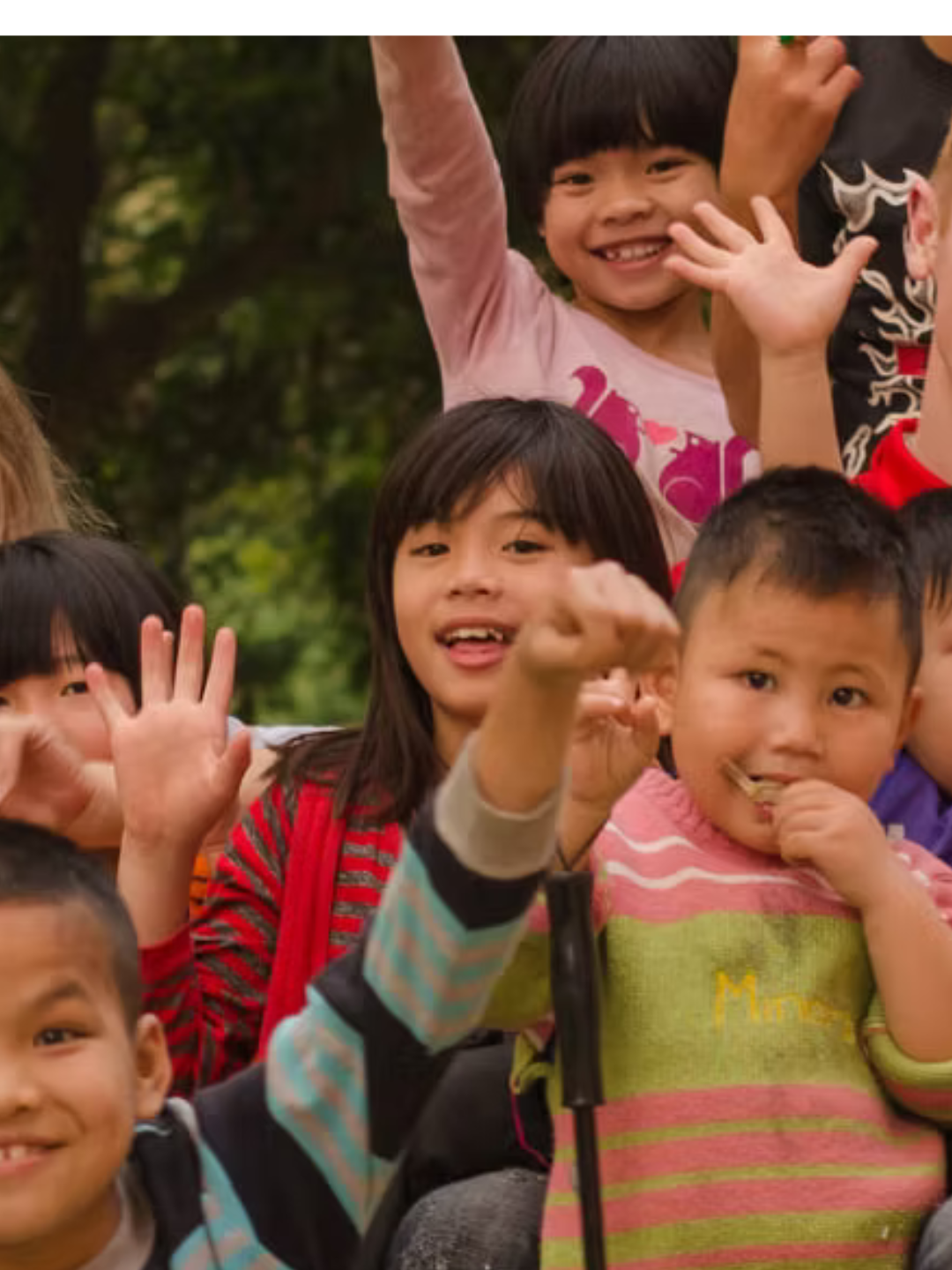 A group of joyful children waving and smiling at the camera outdoors, part of The Collective's charity orphan project.