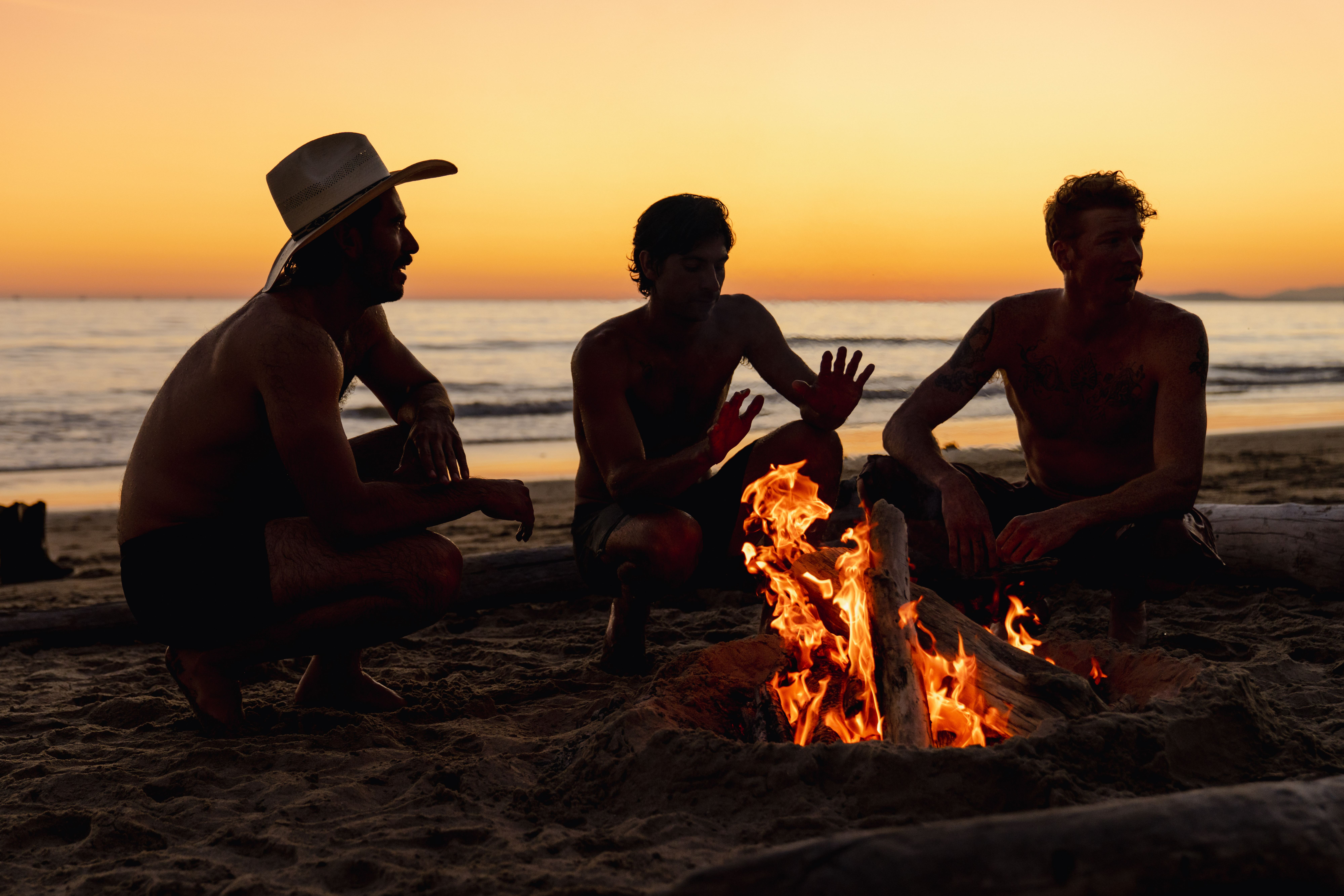 Three men silhouetted around a driftwood bonfire on the beach at sunset